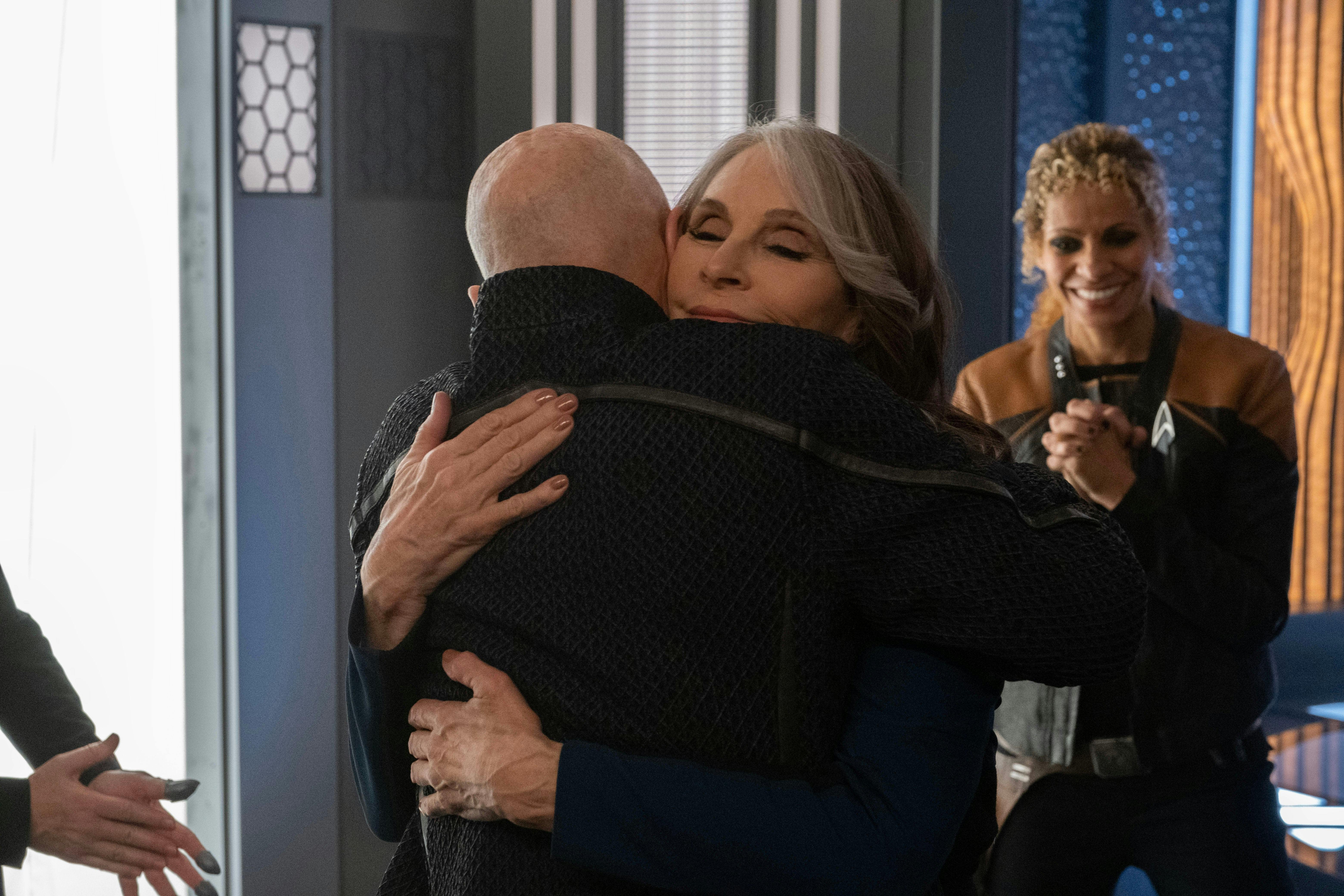Star Trek: Picard BTS still - in the transporter room, Patrick Stewart and Gates McFadden hug as Michelle Hurd smiles and claps behind them