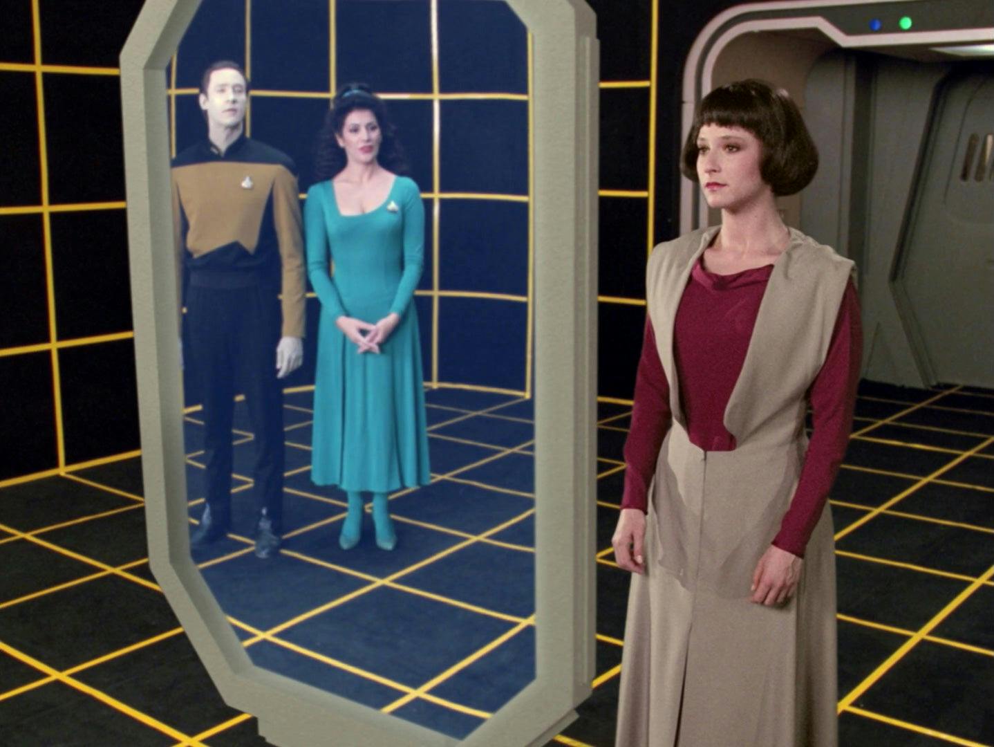 Lal stands in front of a screen, looking at her appearance. Data and Troi can be seen through the screen watching her as she adjusts her appearance.
