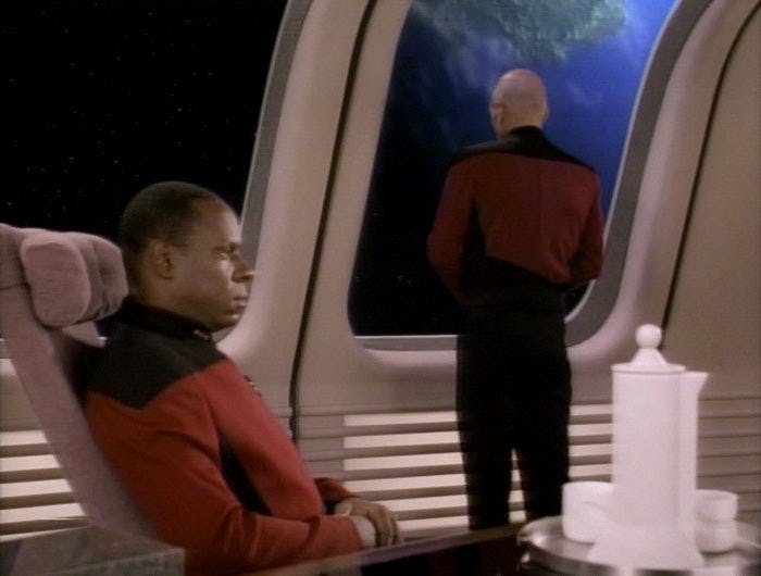 Sisko sits in a conference room onboard the Enterprise-D. Picard is facing out a window, looking away from him.