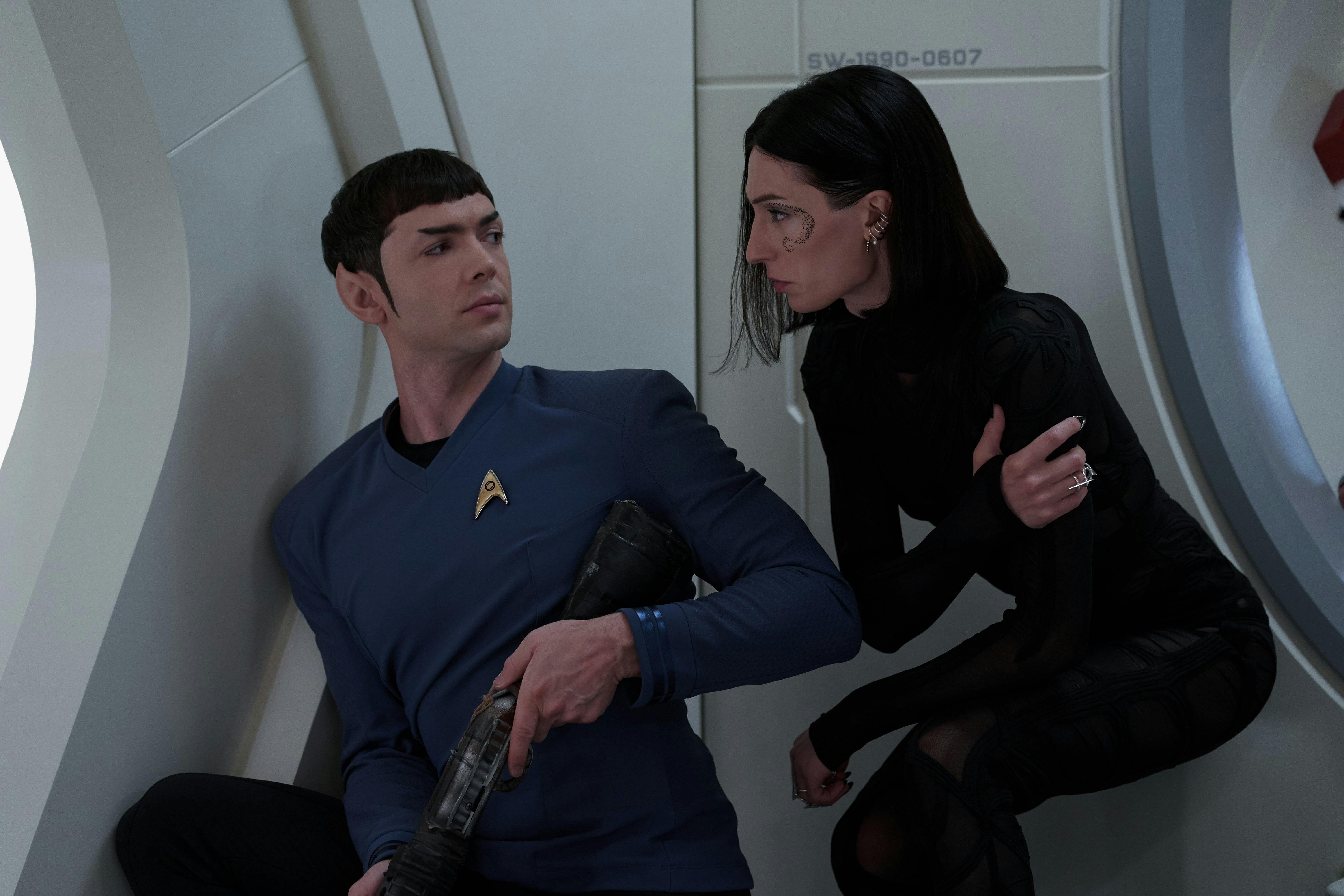 Spock (Strange New Worlds) and a woman with short black hair look at each other. Spock is holding a phaser rifle, and the woman is holding her arm as if she is injured.