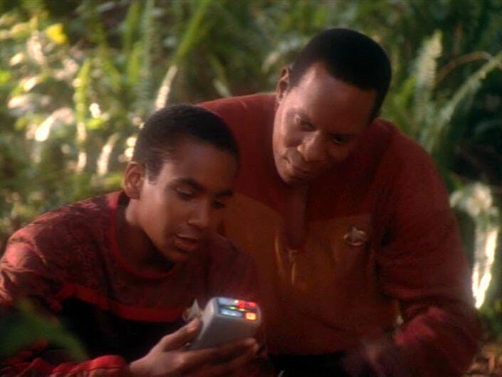 Captain Sisko (Avery Brooks) looks over Jake's (Cirroc Lofton) shoulder as Jake looks at a tricorder.