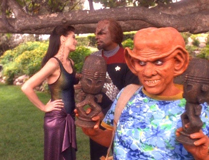 Jadzia Dax, in casual wear, tilts her head with her hands on her hips, knowingly glances at Worf who is in his Starfleet uniform while Quark stands in front of them in casual beach wear holding two horga’hn statues in 'Let He Who is Without Sin...'