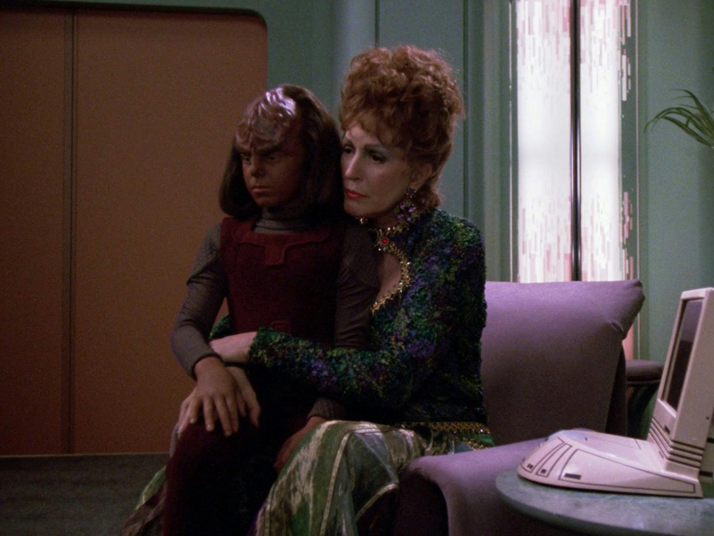 Lwaxana Troi comforts Alexander Rozhenko who sits on her lap while they both of solemn expressions on their faces in 'Cost of Living'