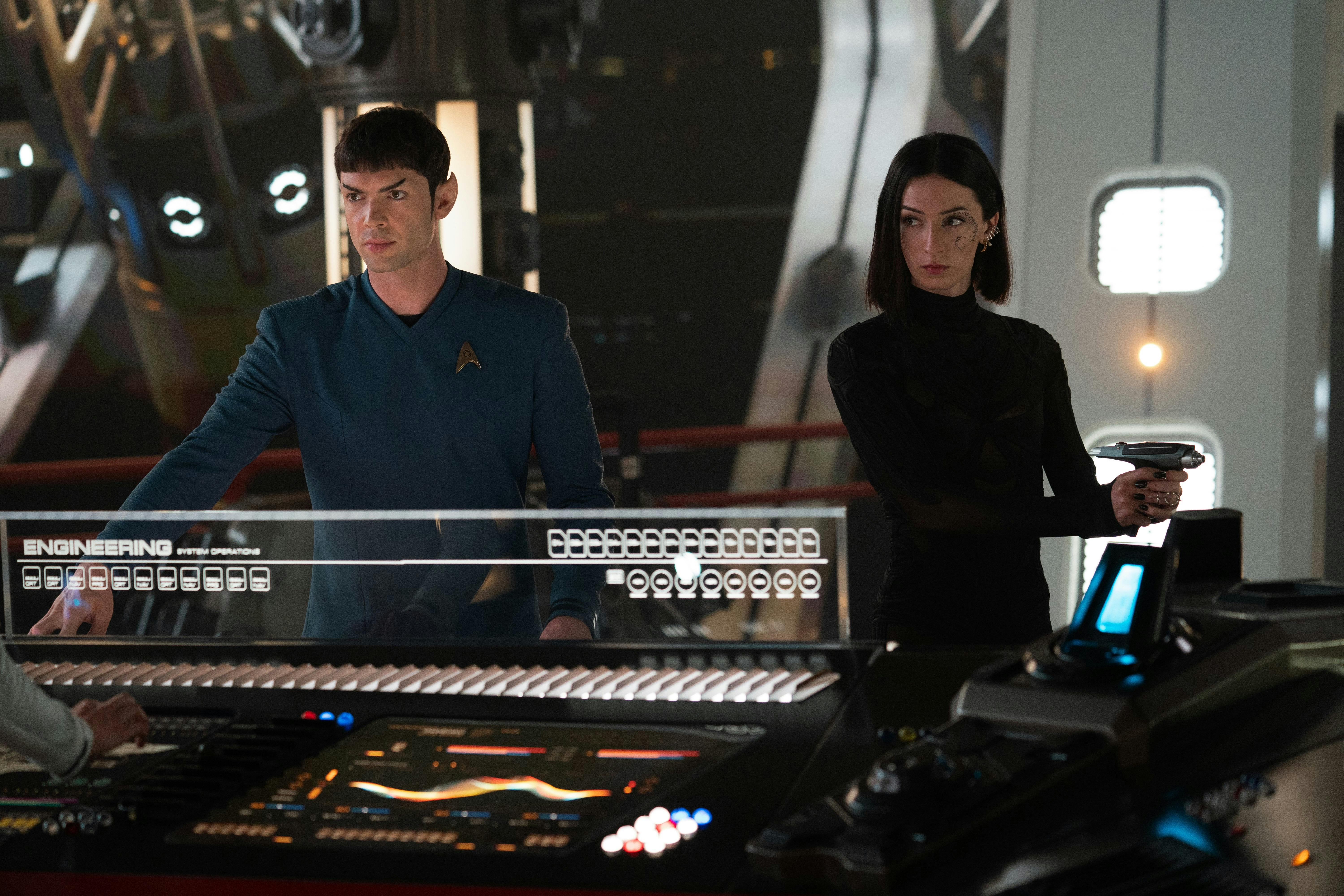 Spock (Ethan Peck) stands next to a console as Dr. Aspen (Jesse James Keitel) stands nearby, holding a phaser.
