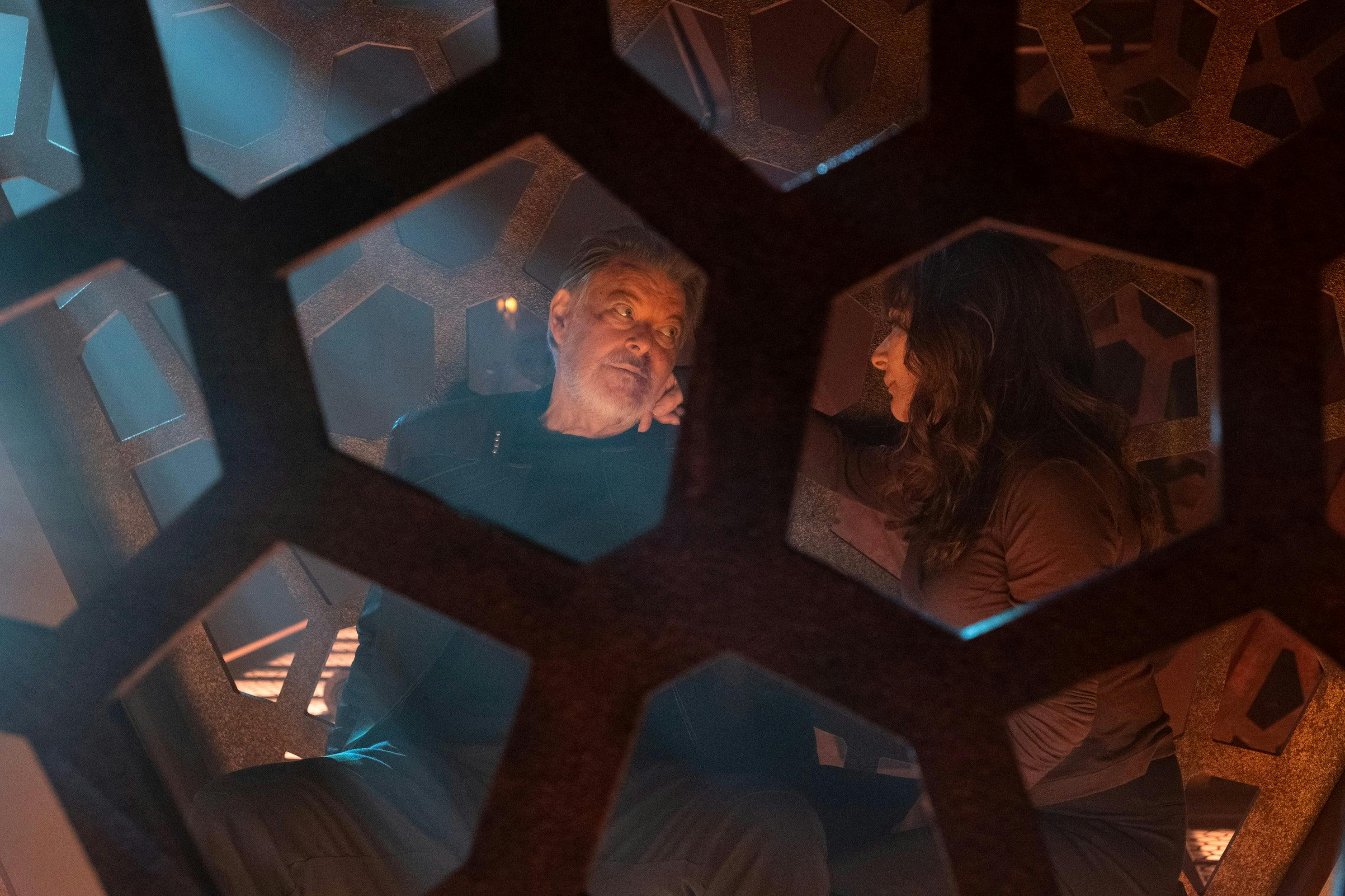 A still of Will Riker and Deanna Troi in a Shrike cell sitting side-by-side looking over at each other as Deanna places her hand on Will's cheek