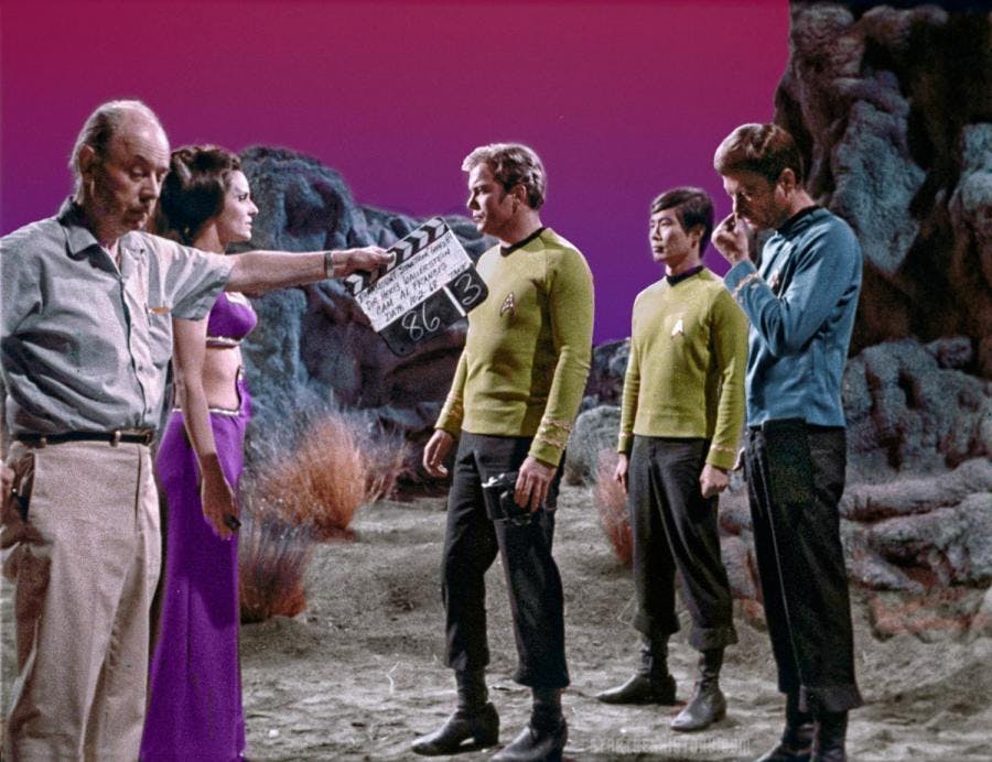 A production still on the set of a surface of a planet as William Shatner, DeForest Kelley, and George Takei standing across from Lee Meriweather as a production member holds a clapboard with details of 'That Which Survives'