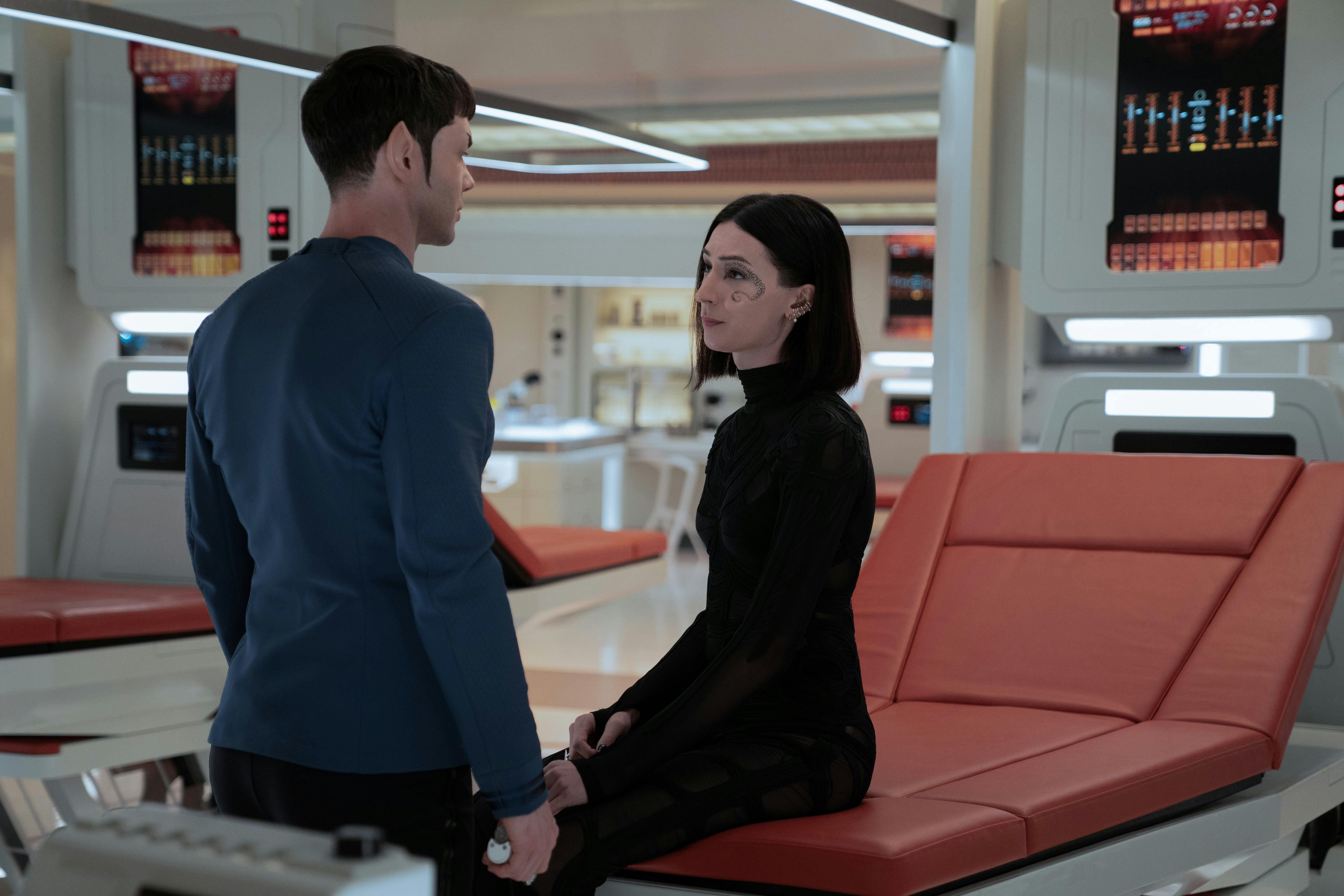 Spock (Strange New Worlds) stands with his back to the camera. He is in medbay, talking to a woman wearing all black with short hair.