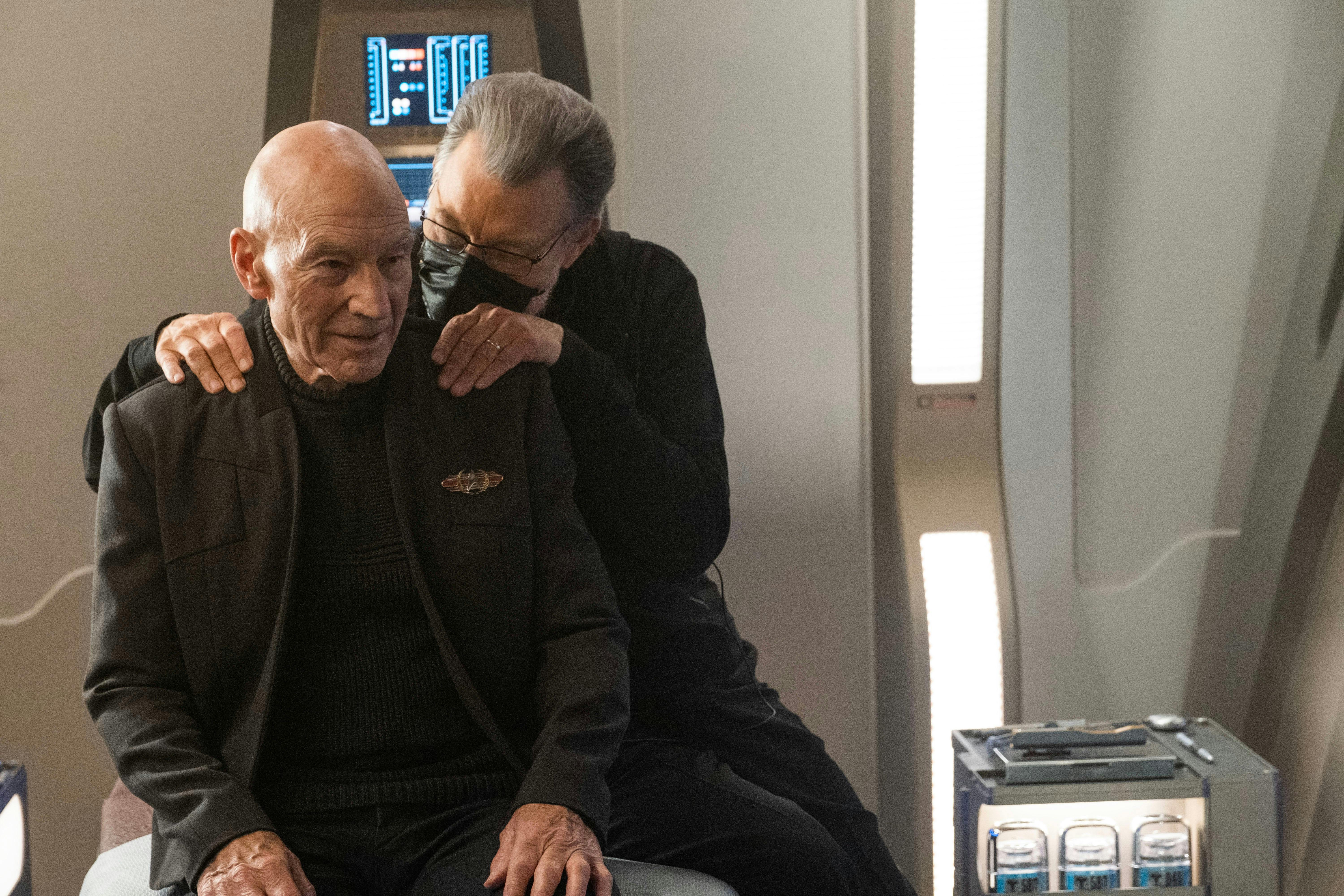 Star Trek: Picard BTS still - in Sickbay, Jonathan Frakes sits next to Patrick Stewart with his hands on his shoulder
