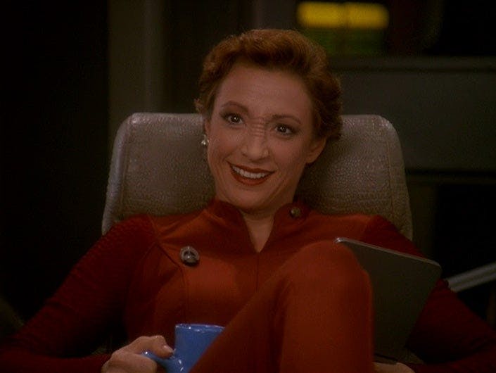 Kira Nerys leans back cozy in her chair, smiling, while holding a mug of raktajino in 'Crossfire'