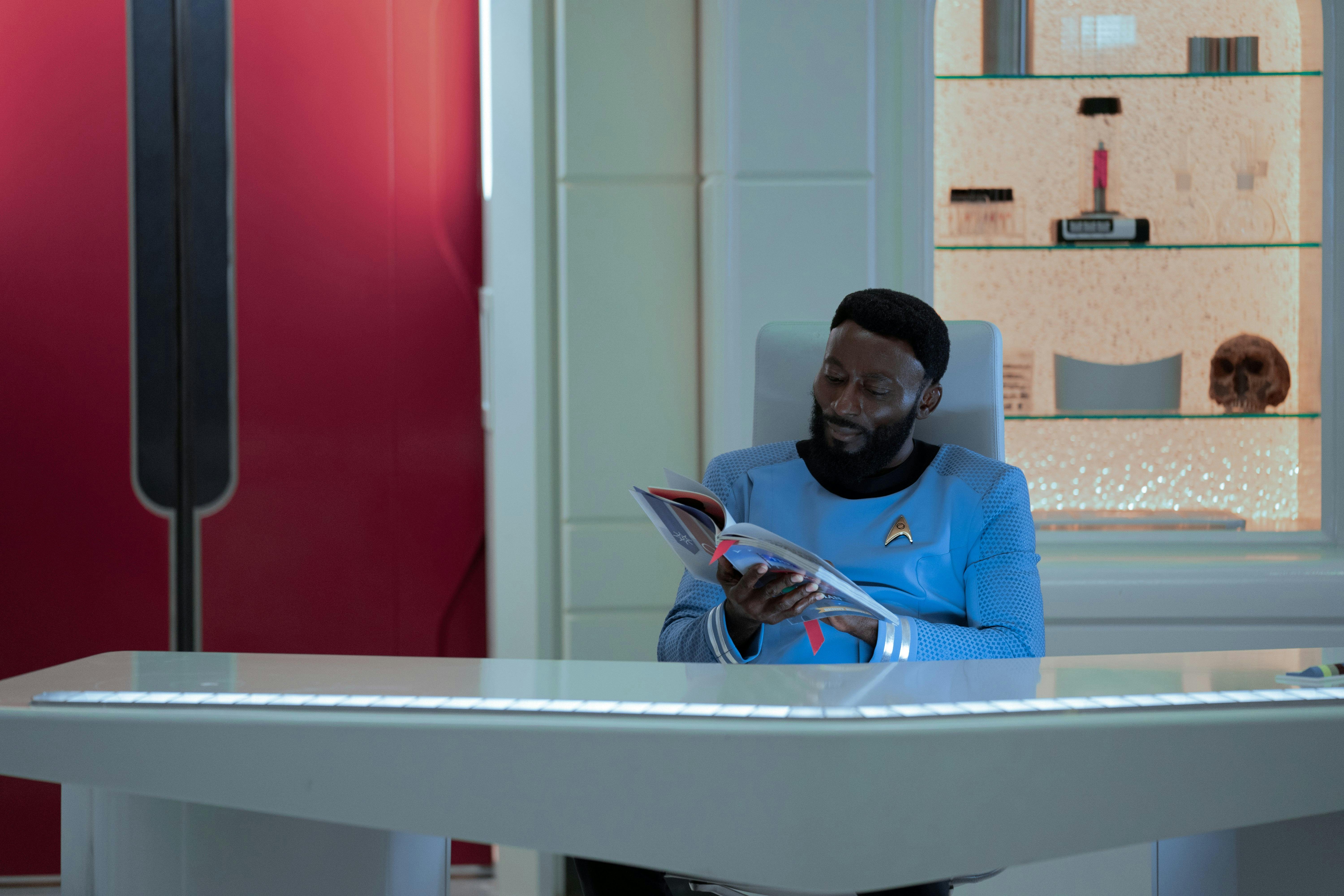 Dr. M'Benga (Babs Olusanmokun) reads a book at his desk.