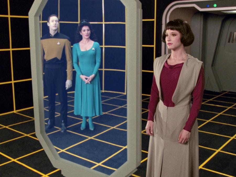 Lal stands in front of a screen, looking at her appearance. Data and Troi can be seen through the screen watching her as she adjusts her appearance.