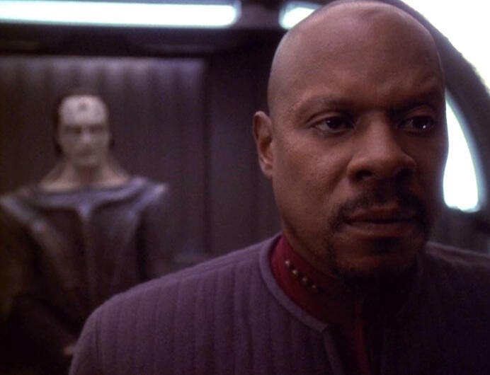 Captain Sisko (Avery Brooks) stands in the foreground of the image. Behind him, out of focus, Gul Dukat is looking at him.