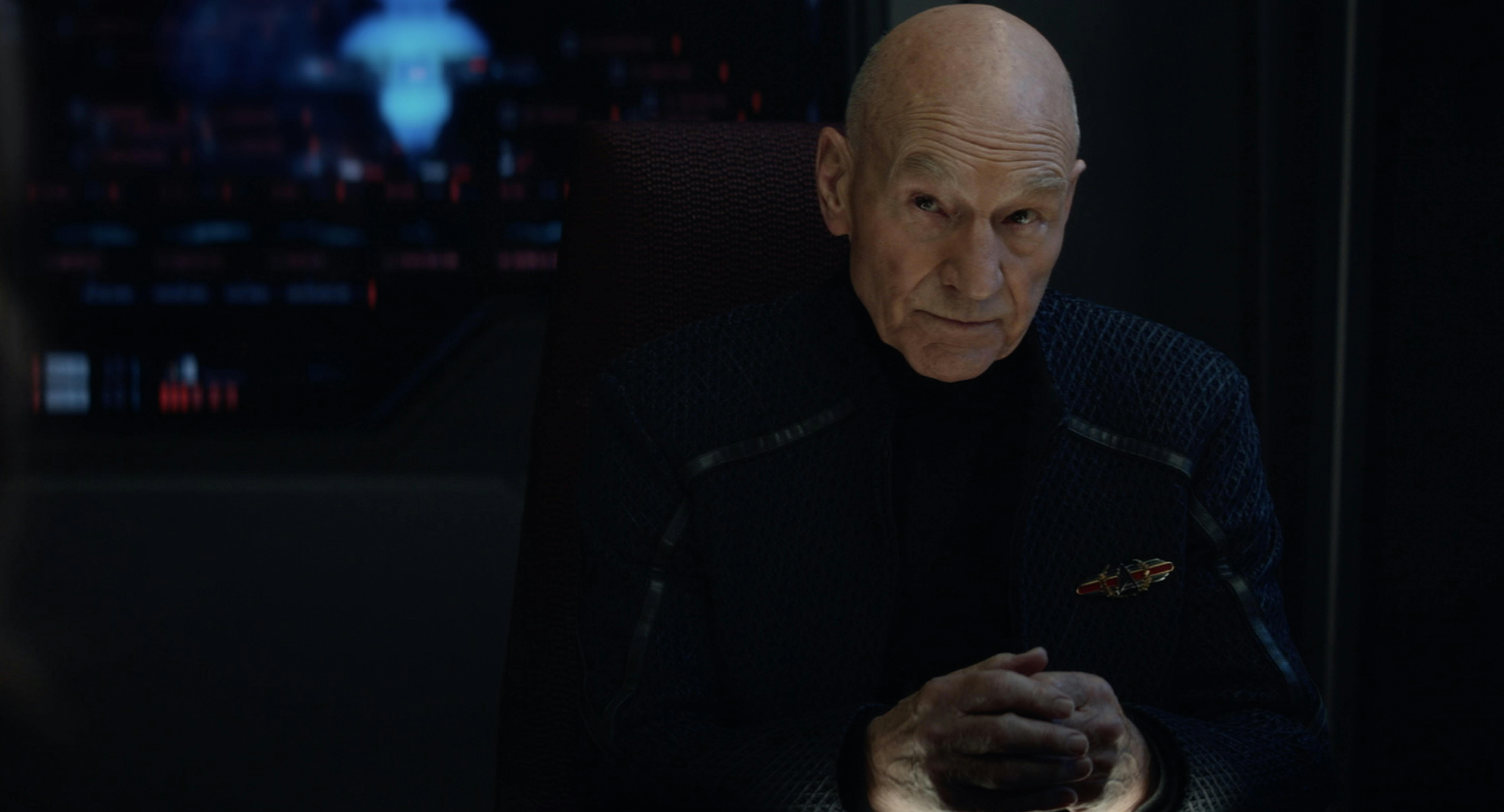 Admiral Jean-Luc Picard sits with his head tilted down but gaze looking up with his hands folded in front of him