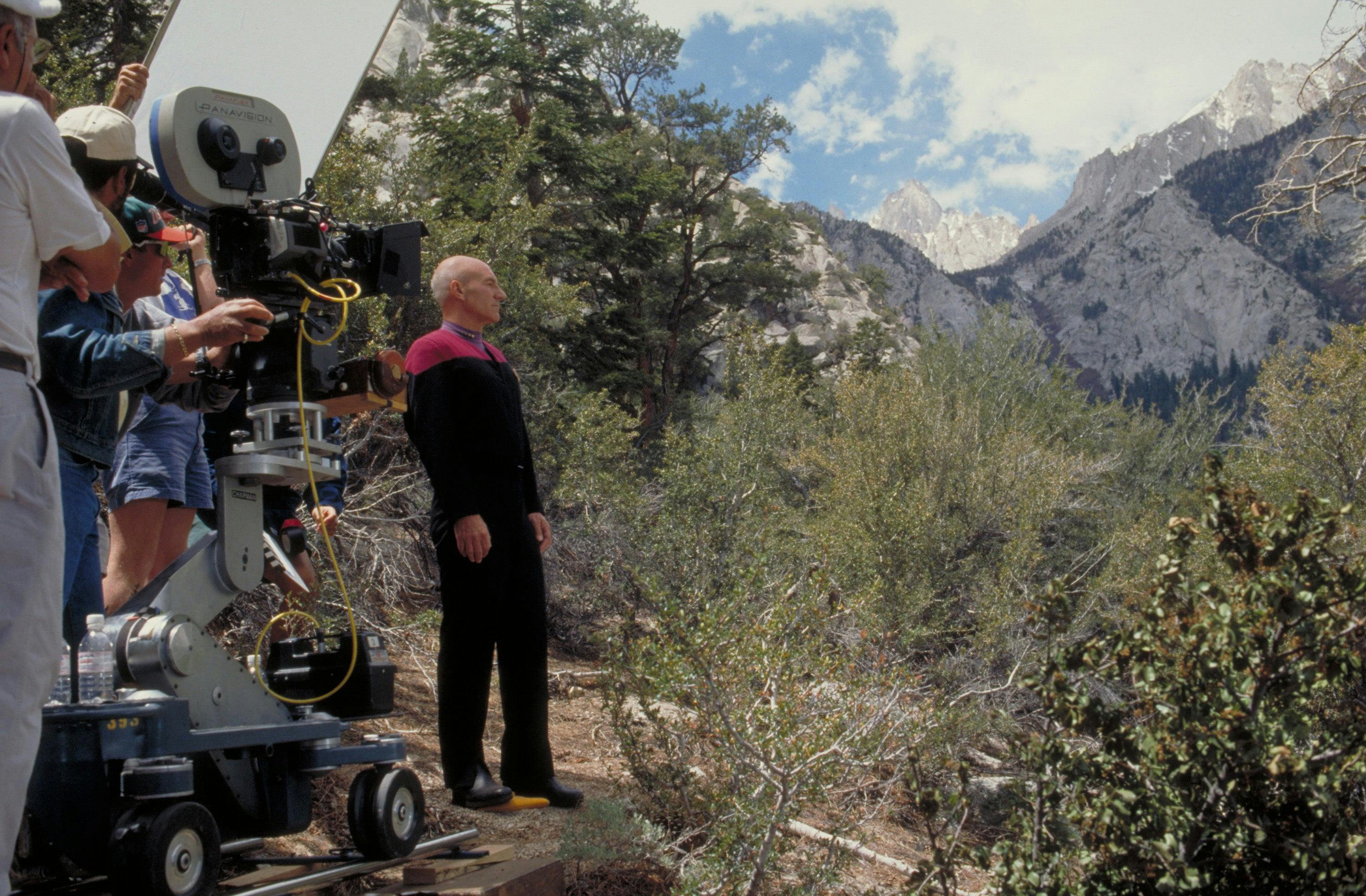 Patrick Stewart, in Starfleet uniform, looking off into the landscape of trees and mountains as a camera and crew capture the moment on set of Star Trek Generations