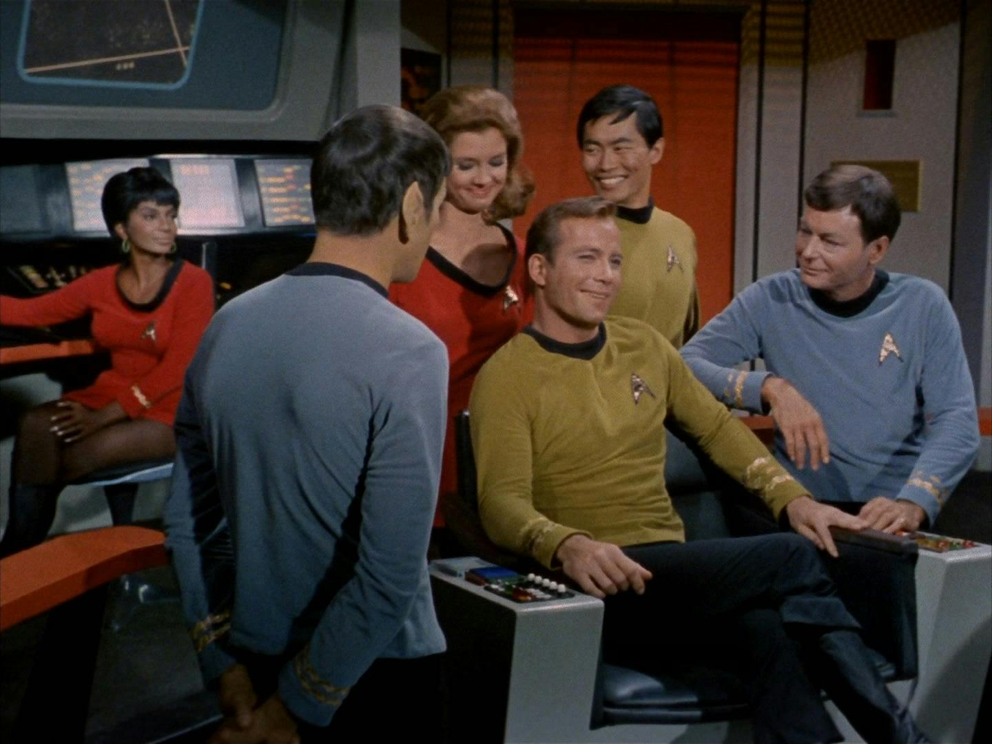 Captain Kirk sits on the bridge in the captain's chair. He is surrounded by his crew and is laughing.