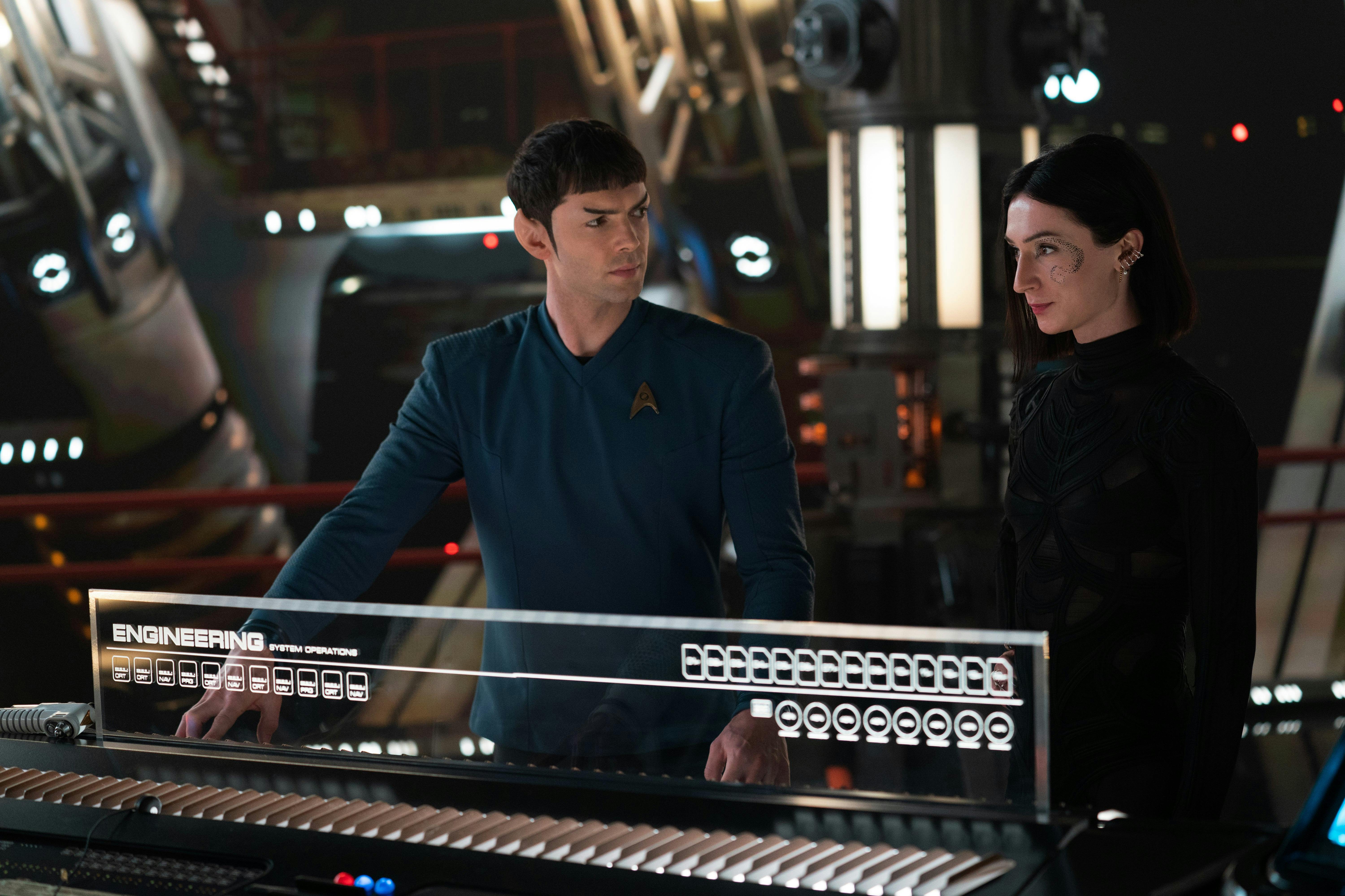 Spock (Ethan Peck) and Dr. Aspen (Jesse James Keitel) stand next to a console, looking at each other.