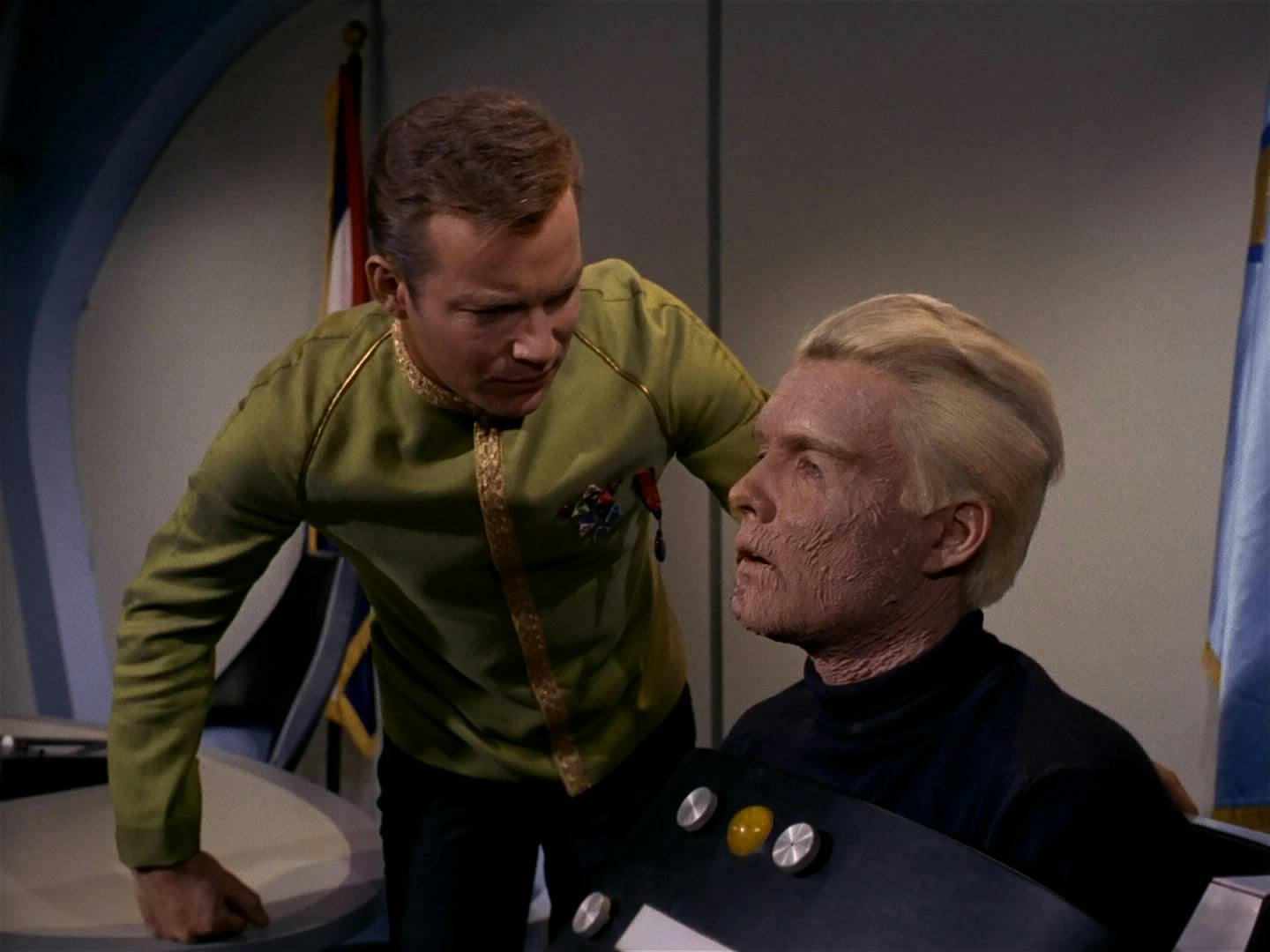 Captain Kirk stands next to Captain Pike, who is disabled after his accident and is in a futuristic wheelchair.