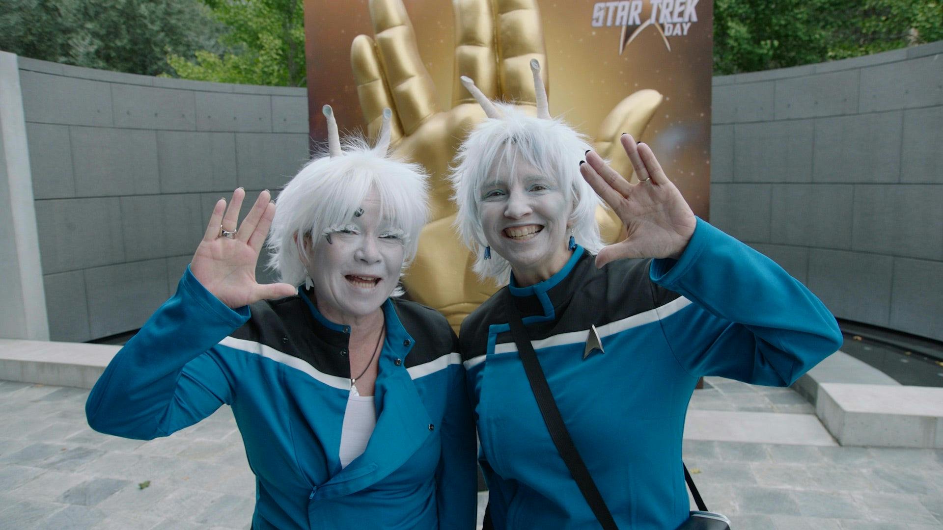 Two fans dressed as Aenar pose in front of a Vulcan salute statue. They are wearing blue Lower Decks uniforms and are giving the camera the Vulcan salute.