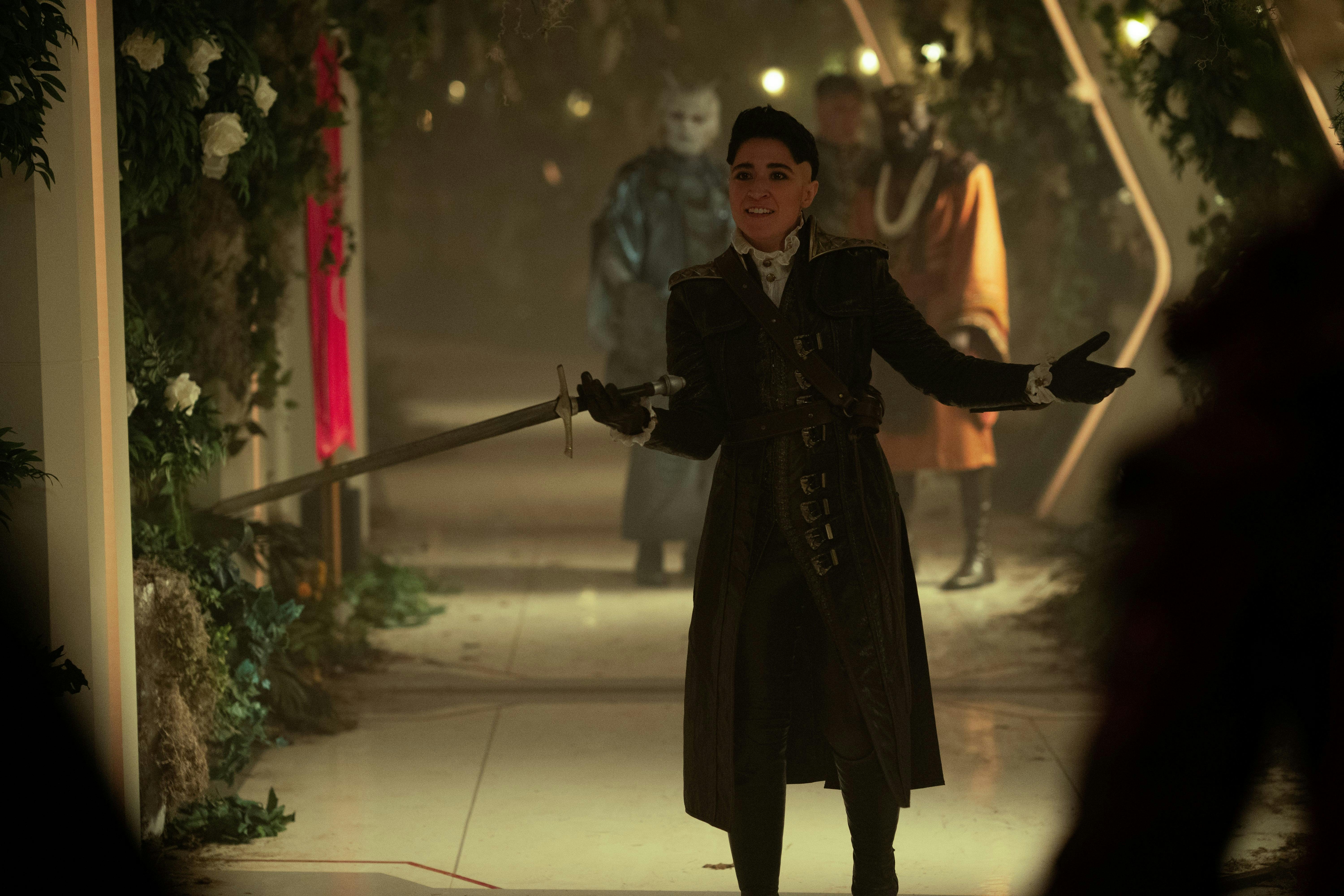 Ortegas (Melissa Navia), holding a sword and wearing medieval clothes, stands in the middle of the Enterprise's hallway. The hallway is overgrown with leaves. There is a small group standing behind her.