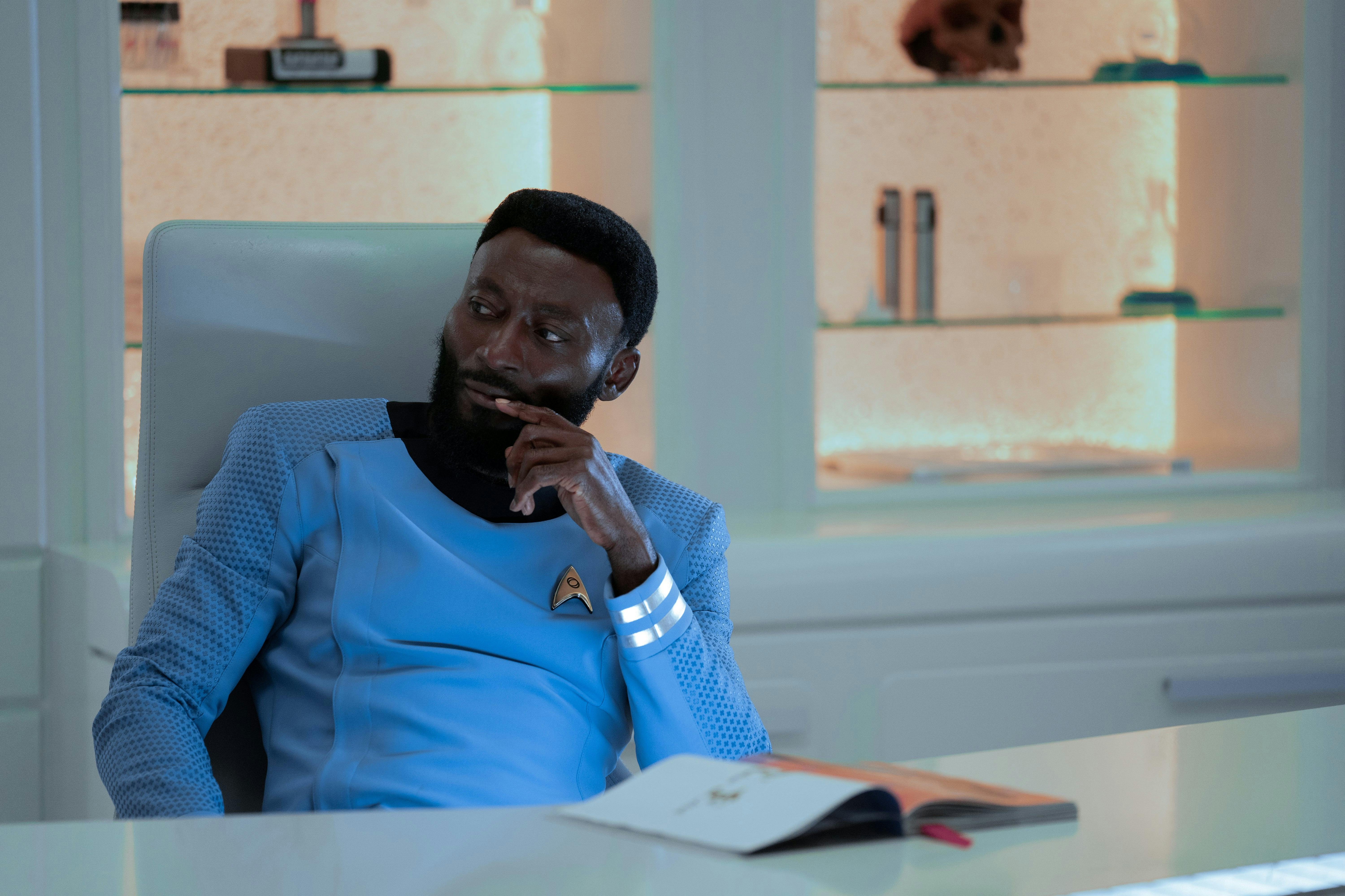 Dr. M'Benga (Babs Olusanmokun) sits at his desk, looking thoughtful.