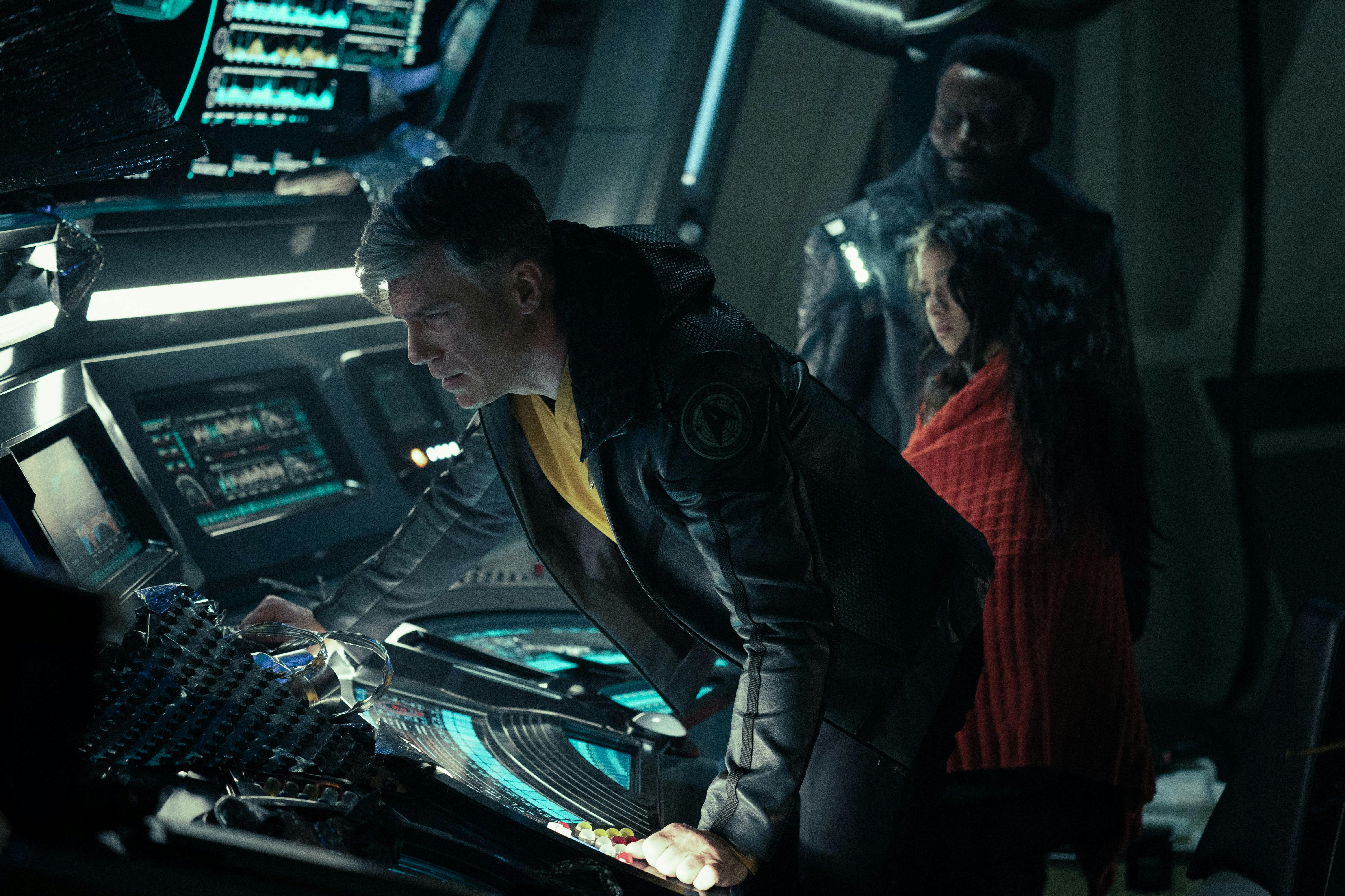 Captain Pike (Anson Mount) leans over a console. Next to him, a young woman wearing red and Dr. M'Benga (Babs Olusanmokun) stand watching him.