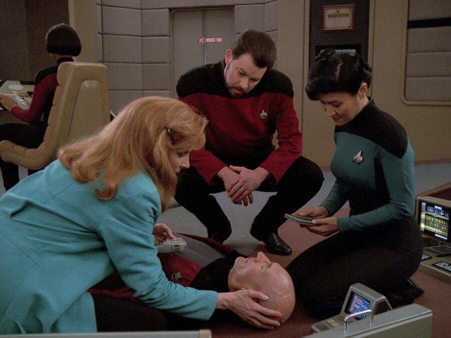 Captain Picard lays on the bridge of the Enterprise-D while Dr. Crusher, Nurse Alyssa Ogawa, and Will Riker crowd and monitor him in 'Inner Light'