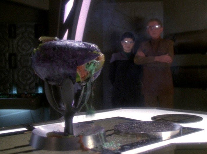 Ezri and Odo, with safety goggles on, witness O'Brien's demonstration of the TR-116 on a melon