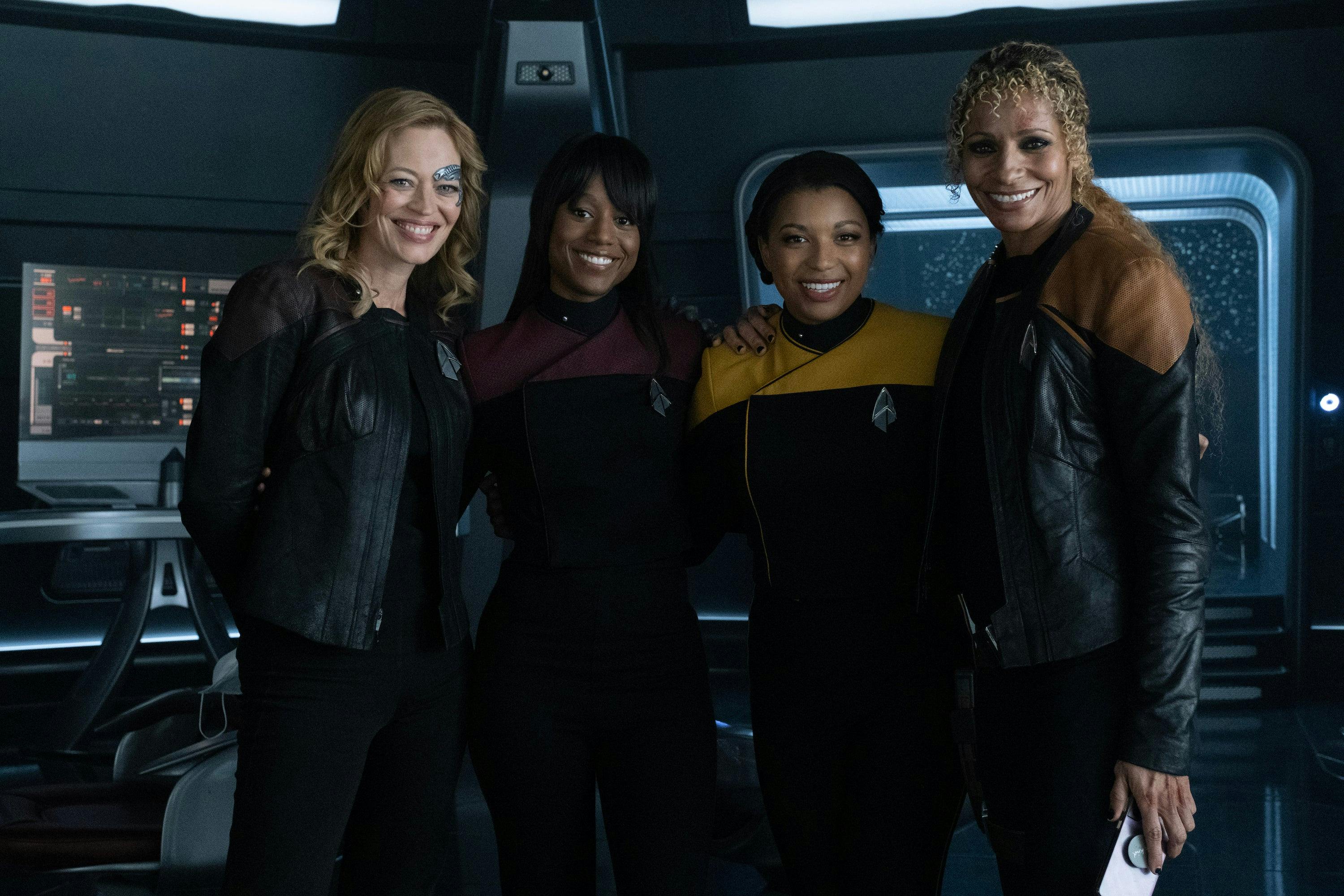 Star Trek: Picard BTS still - Jeri Ryan, Ashlei Sharpe Chestnut, Mica Burton, and Michelle Hurd smile and stand on the bridge of the Titan set