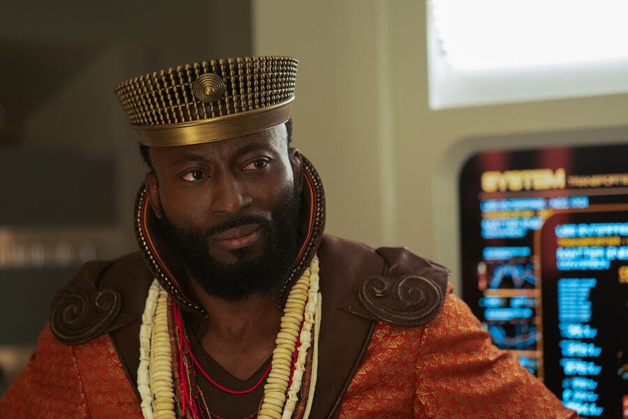 Dr. M'Benga (Babs Olusanmokun), wearing a crown and robes, stands near a console.