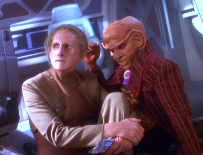 Odo sits on the floor wrapping his arm over his left leg as Quark sits right beside him in 'The Ascent'