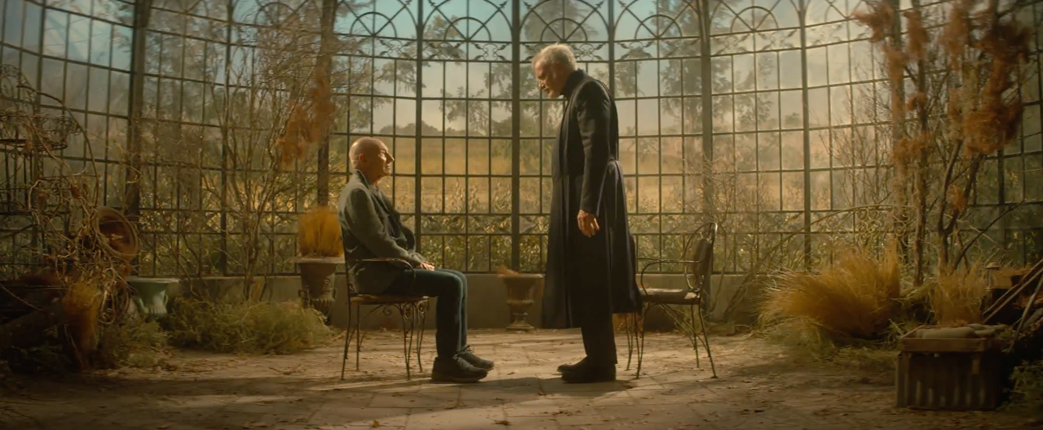 Jean-Luc sitting while Q stands in front of him in the greenhouse at the chateau. 