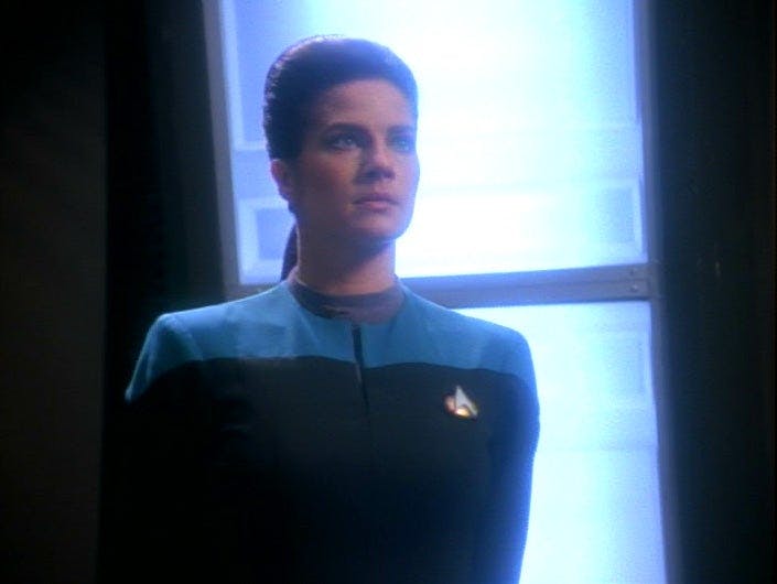 Jadzia Dax stands against a bright blue light. She is wearing a black and blue uniform.