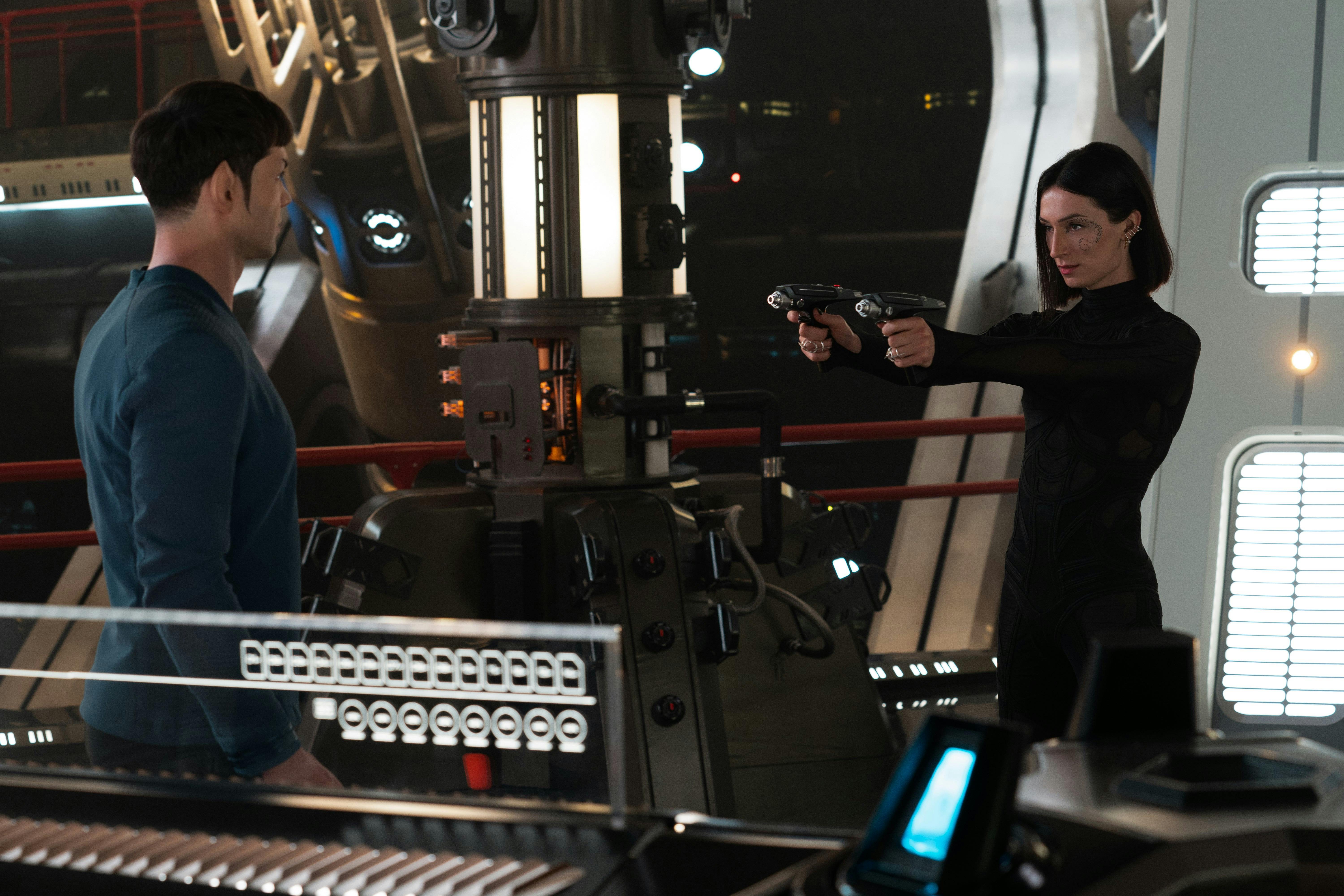 Dr. Aspen (Jesse James Keitel) stands with two phasers aimed at Spock (Ethan Peck), who's back is to the camera.