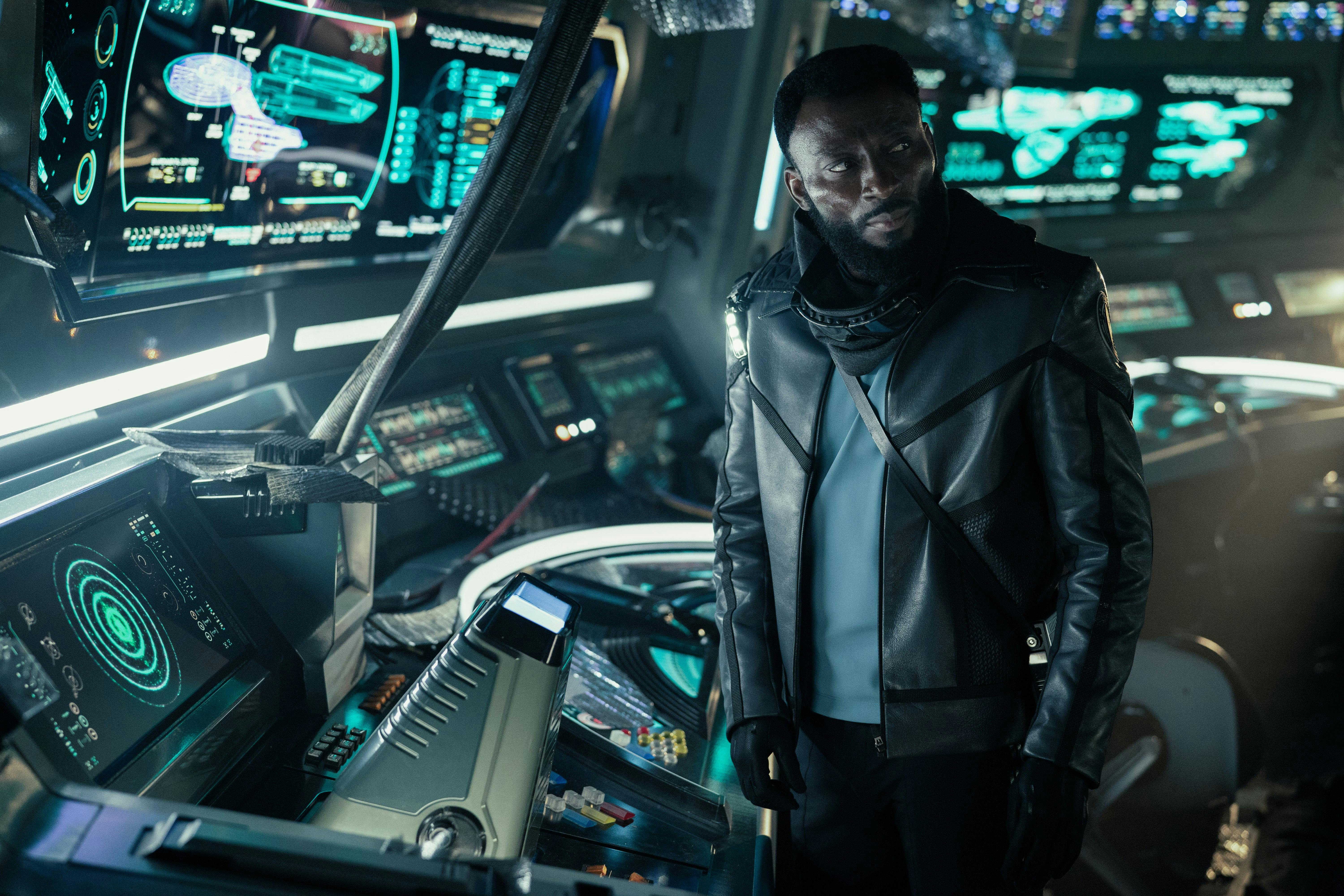 Dr. M'Benga (Babs Olusanmokun) stands near a console on the Enterprise's bridge. He is wearing a jacket over his uniform.