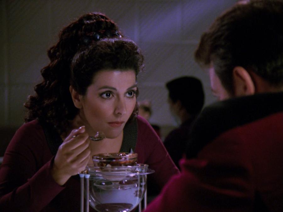 Deanna Troi leans forward while eating a chocolate sundae in Ten Forward. Riker is sitting across from her, with his back to the camera.