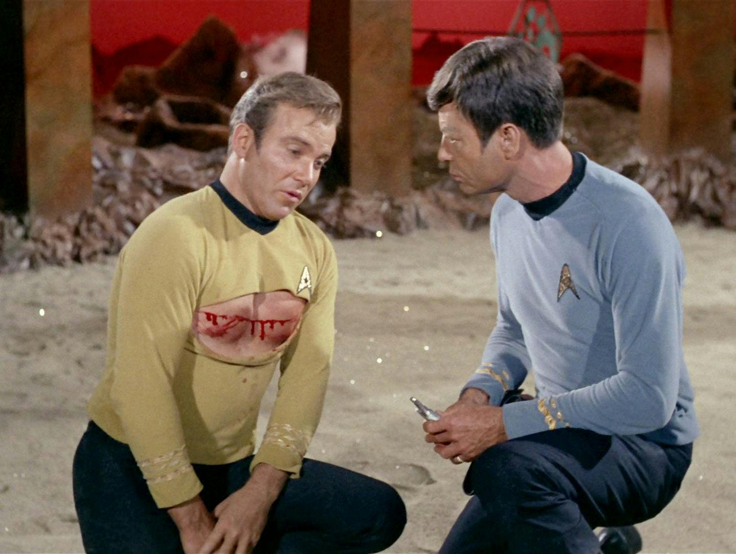 On Vulcan, McCoy crouches beside Captain Kirk who has his uniform ripped and a bloody gash across his chest in 'Amok Time'