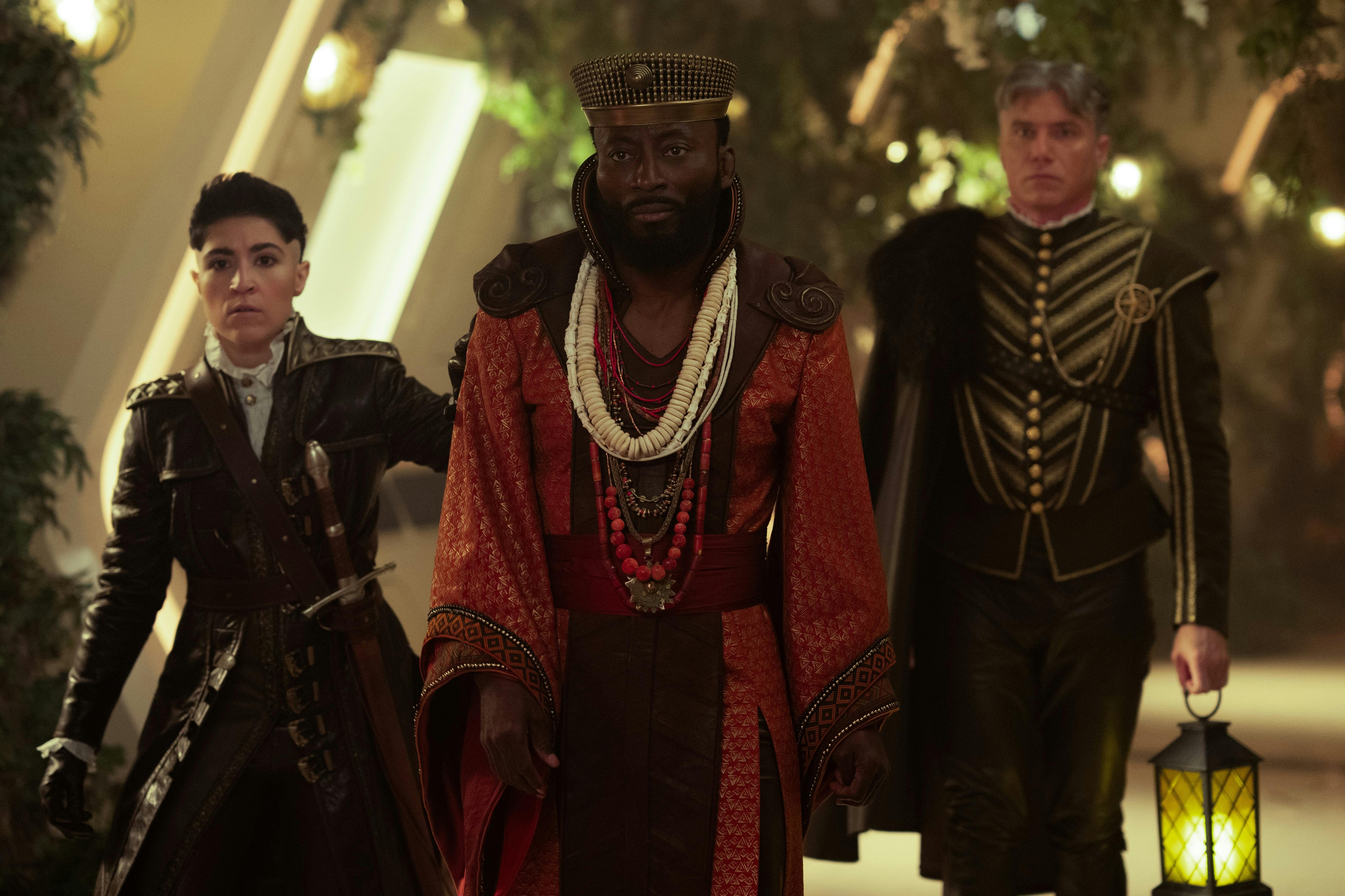 M'Benga (Babs Olusanmokun), wearing robes and a crown, walks down the Enterprise's hallway. Ortegas (Melissa Navia) and Pike (Anson Mount) follow him. Pike holds a lantern, and both characters are dressed in medieval costumes in 'The Elysian Kingdom'