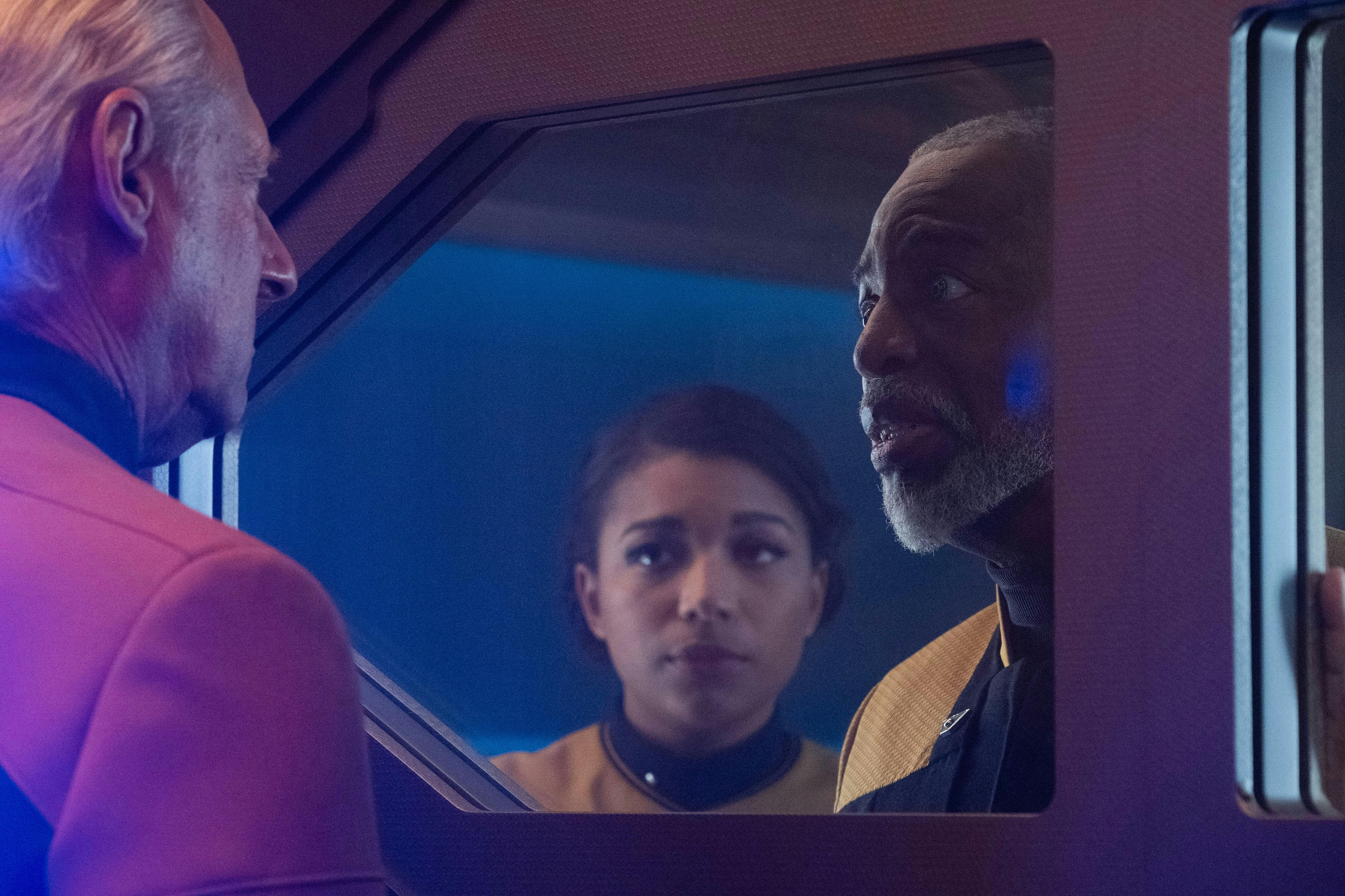 Geordi La Forge looks at Lore behind the Control Room door as a concerned Alandra looks on