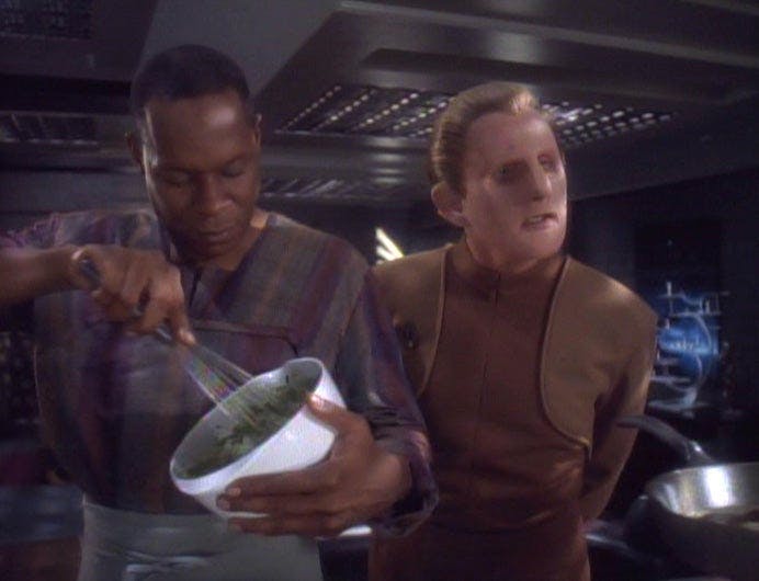 Sisko in civilian attire and an apron mixes a bowl with a whisk as Odo looks over his shoulder at the spread in 'Equilibrium'