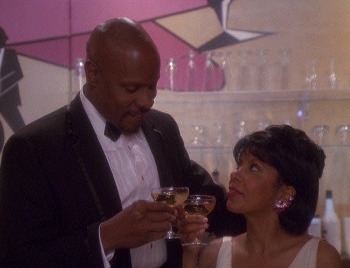Ben Sisko raises his champagne glasses to a seated Kasidy Yates' glass while they're in the holosuite in Star Trek: Deep Space Nine's 'Badda-Bing, Badda Bang'