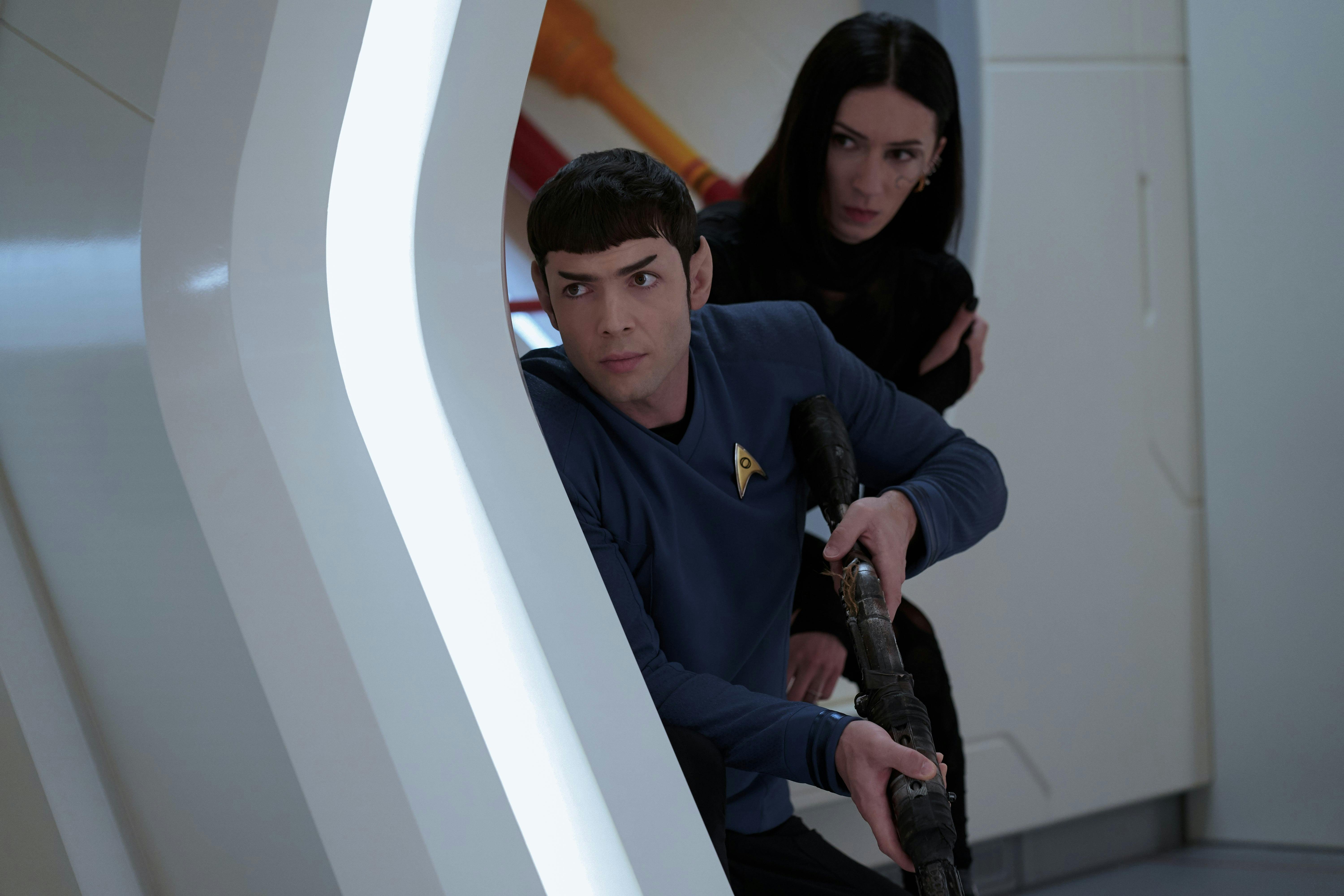 Spock (Strange New Worlds) is sheltered behind a door in the medbay, holding a phaser rifle at the ready. A woman with short black hair is behind him.