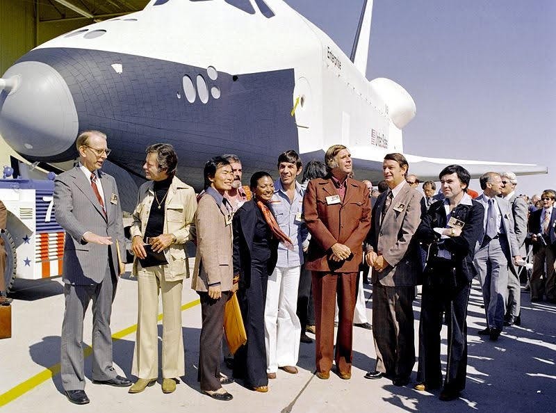Star Trek actors with Gene Roddenberry and NASA administrators stand before a prototype of the space shuttle test vehicle Enterprise in 1976. NASA administrator James Fletcher speaks to DeForest Kelly while George Takei, Nichelle Nichols, Leonard Nimoy, Gene Rodenberry and Walter Koenig take it all in. Star Trek fans had organized a letter-writing campaign to have this early space craft named after their favorite starship.