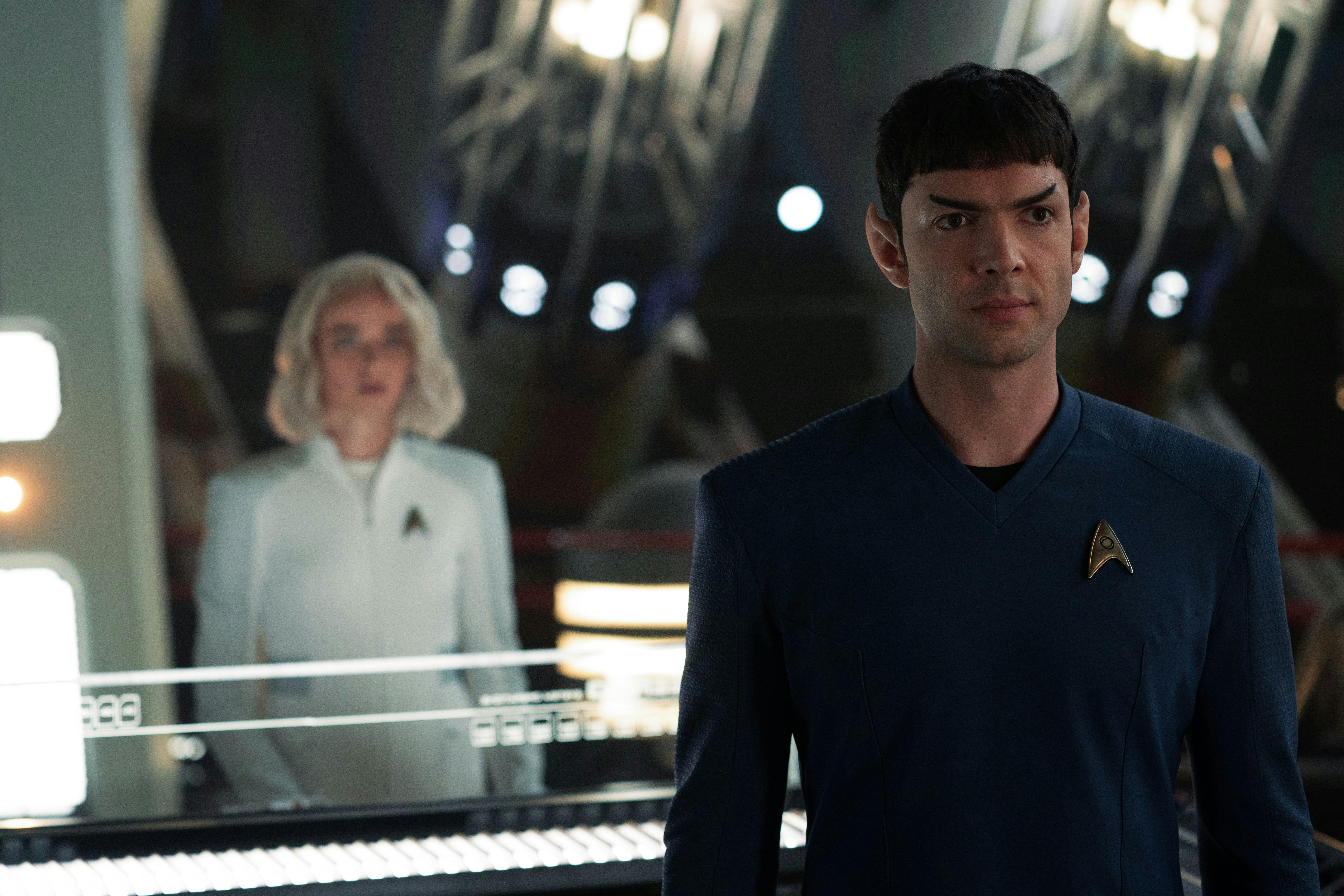 Spock (Strange New Worlds) looks at someone off camera with a neutral expression. To his right and out of focus, Chapel stands a little ways behind him.