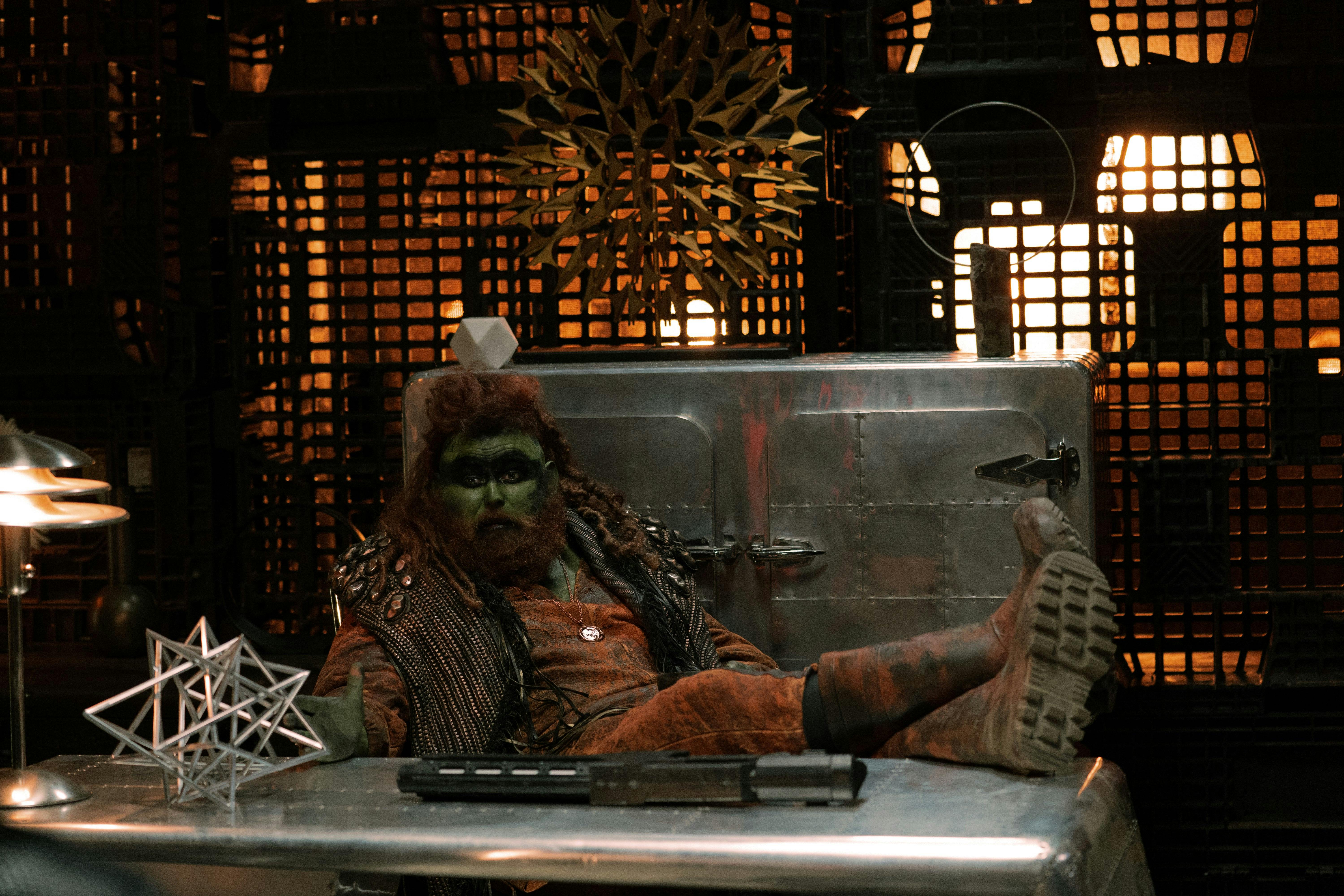 A green alien with red hair and a red beard sits behind a desk with his feet up.
