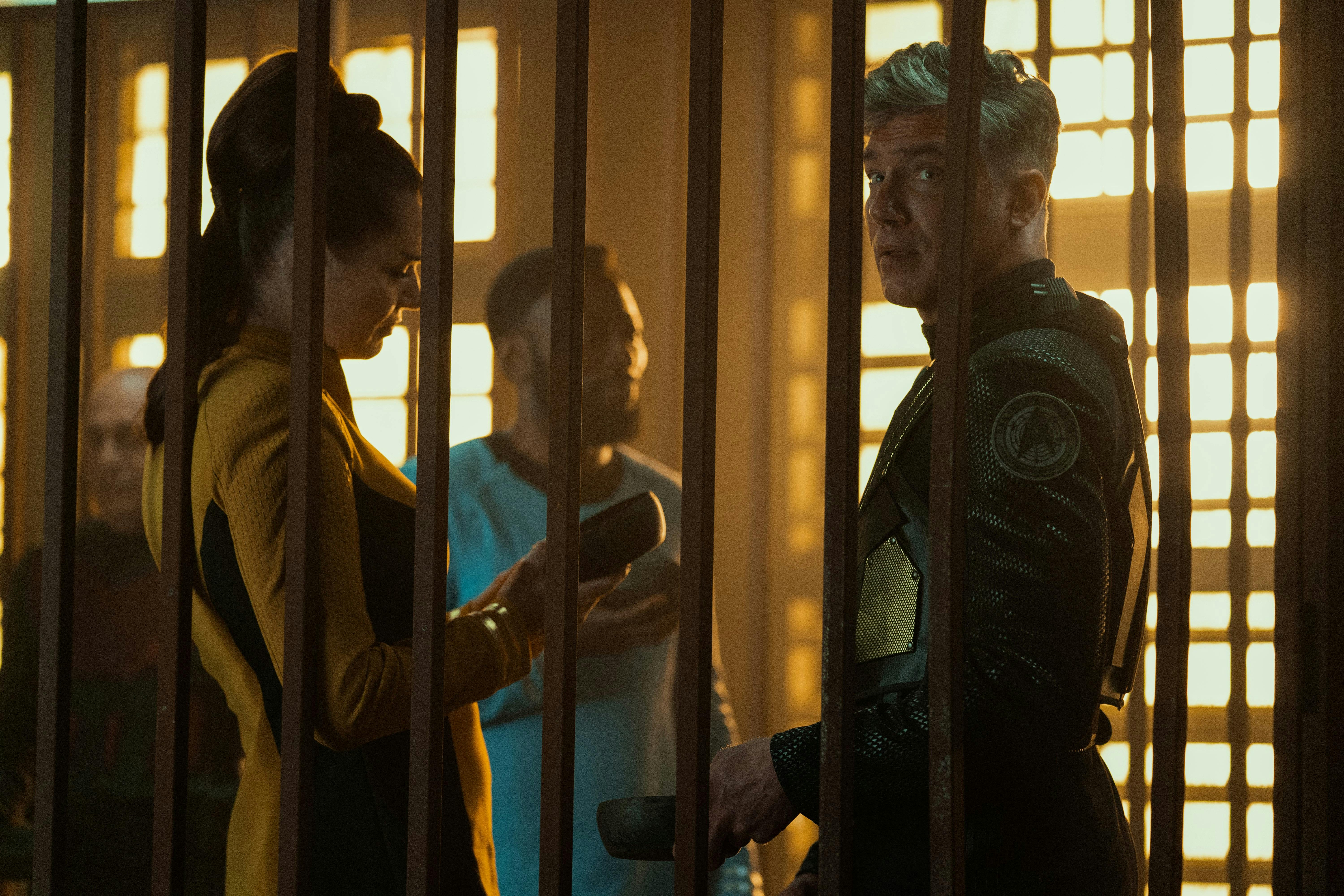 Number One (Rebecca Romijn) and Pike (Anson Mount) confer while being held behind bars. In the background, you can see Dr. M'Benga (Babs Olusanmokun).
