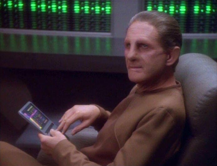Odo sits back in his chair with his PADD in hand while looking up in 'Q-Less'
