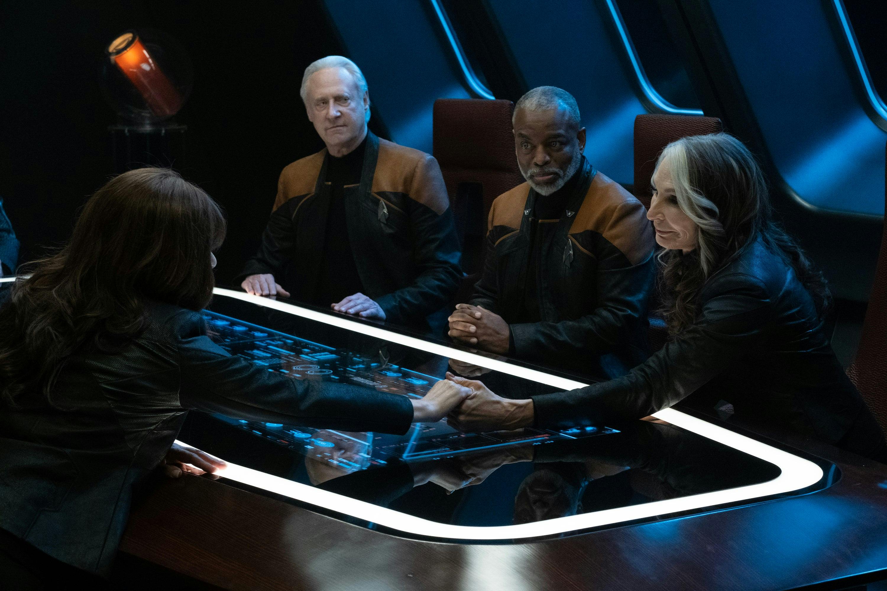 In the Titan's Observation Lounge, a seated Deanna Troi and Beverly Crusher reach across the table holding each other's hands as Data and Geordi La Forge observe