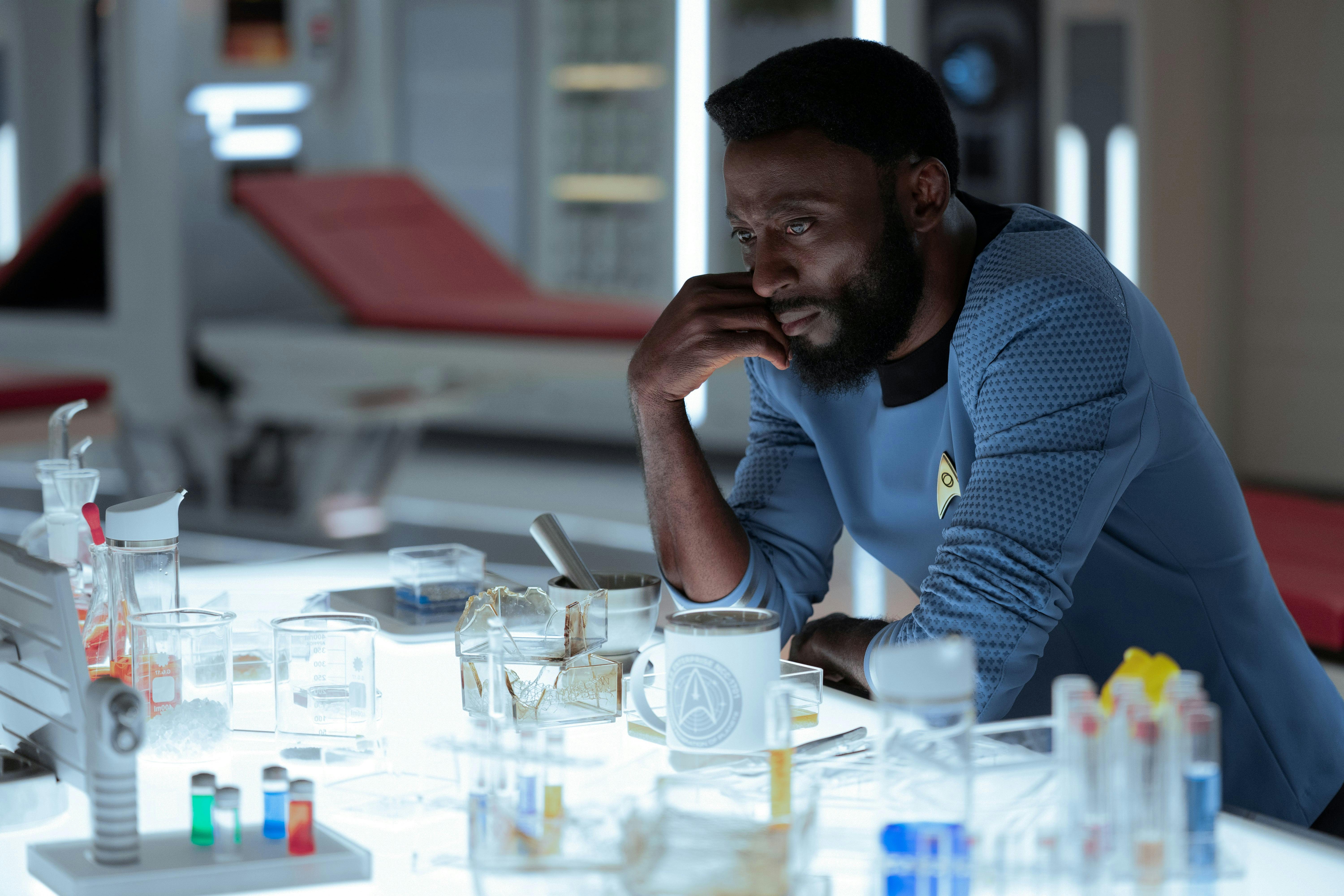 Dr. M'Benga (Babs Olusanmokun) sits at a table in medbay. He studies the equipment in front of him.