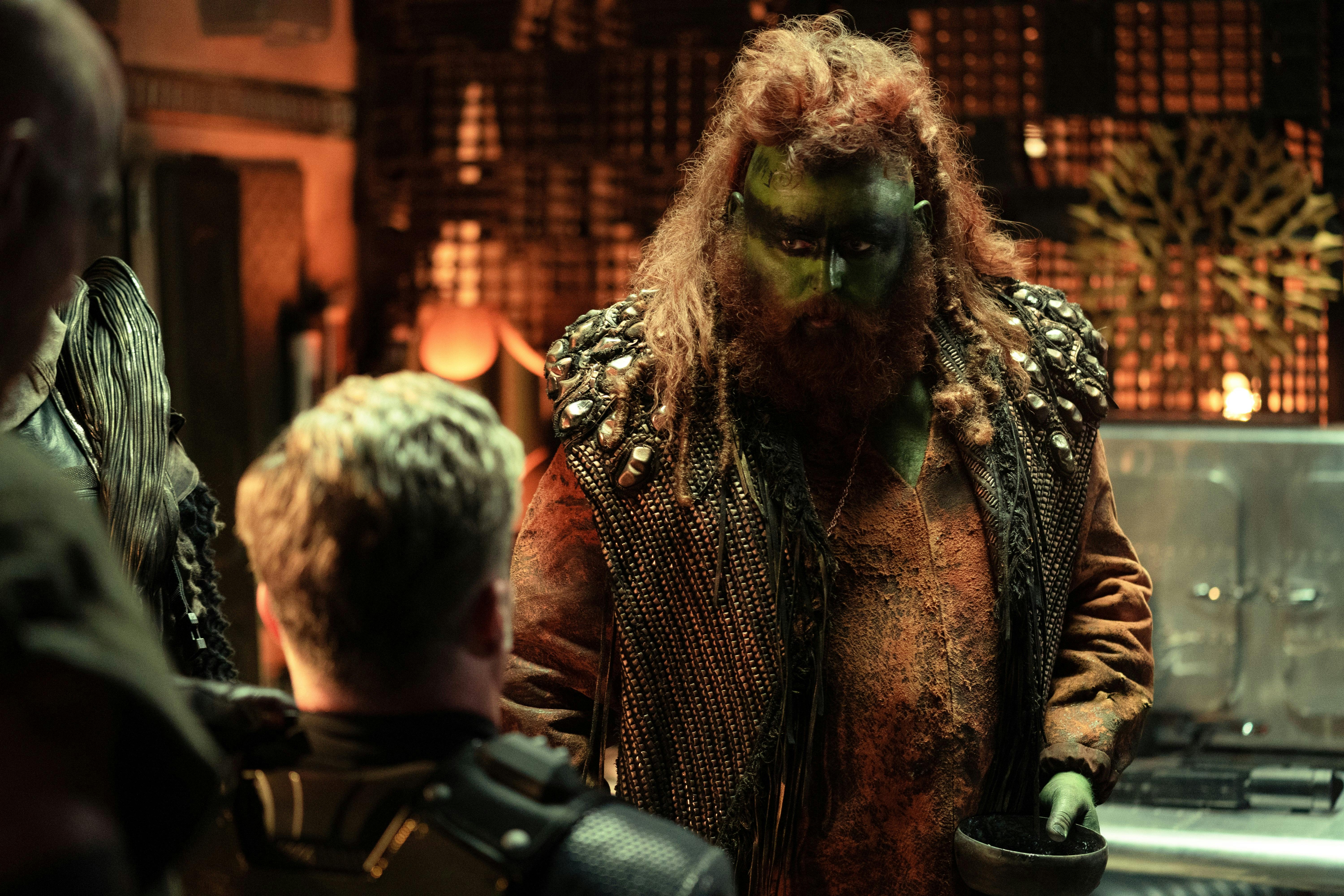 An alien with green skin and orange hair and beard stands in front of a kneeling Pike (Anson Mount). Pike's back is facing the camera.