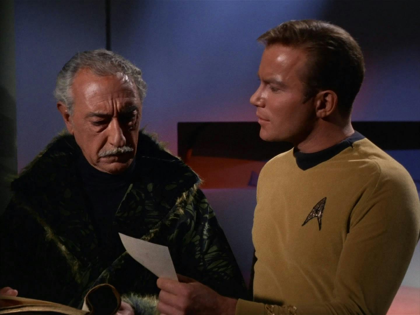 Captain Kirk (The Original Series) stands next to Kodos, and holds out a piece of paper to him.